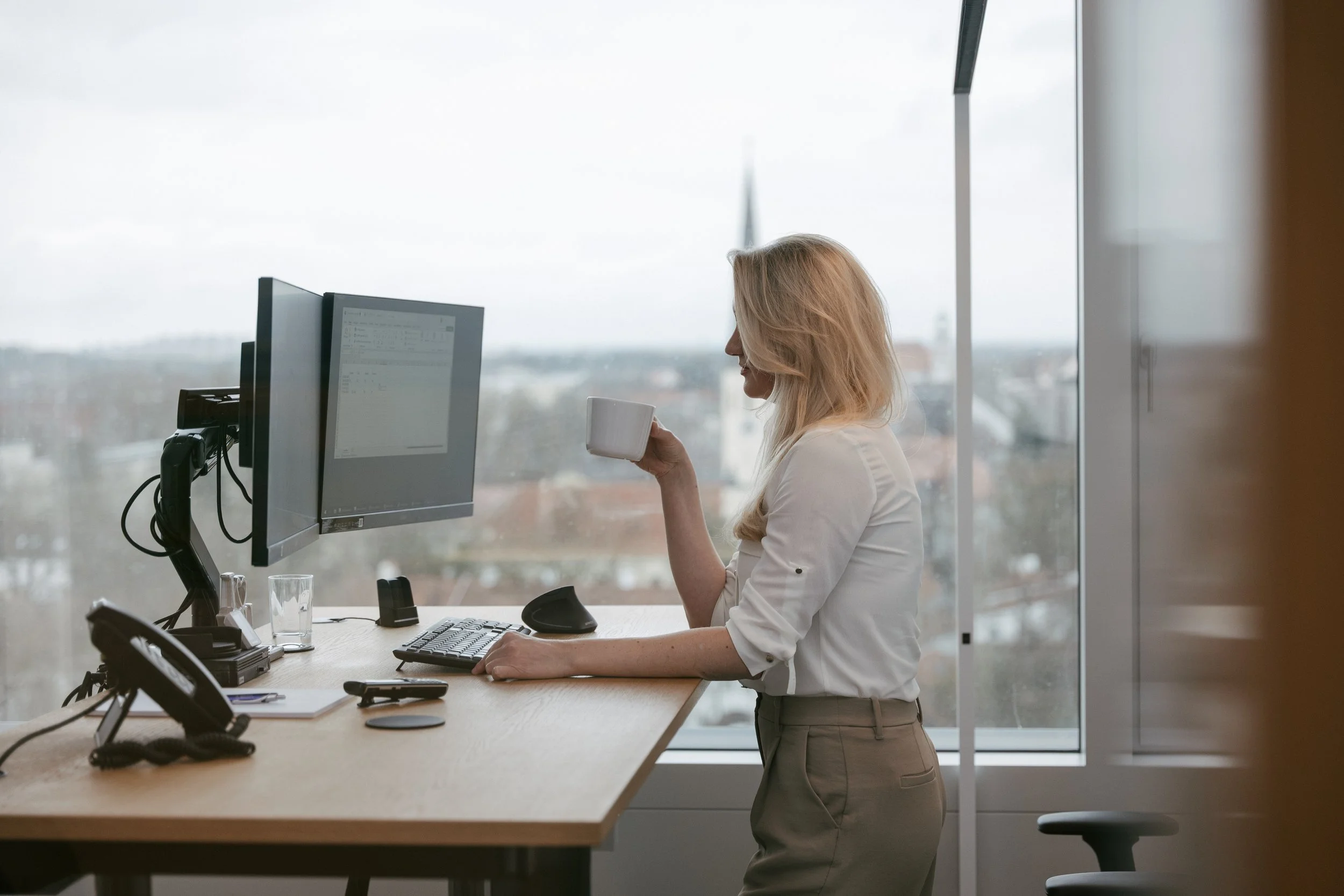 Frau steht in einem Büro am Schreibtisch, trinkt aus einer Tasse und schaut auf einen Computerbildschirm. Draußen ist eine Stadtansicht durch das Fenster sichtbar.