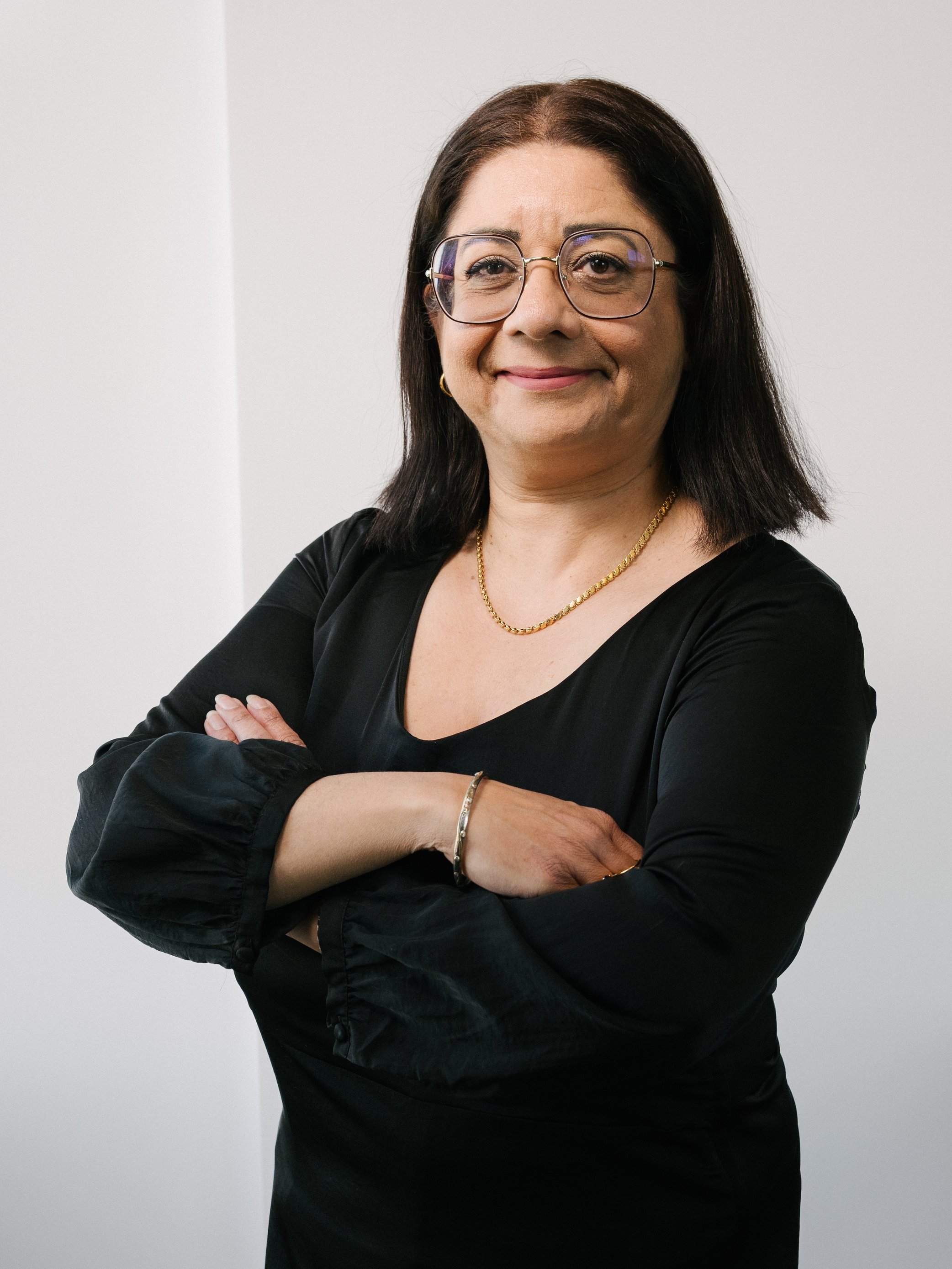 Frau mit Brille, schwarzem Kleid und verschränkten Armen steht vor einem weißen Hintergrund.