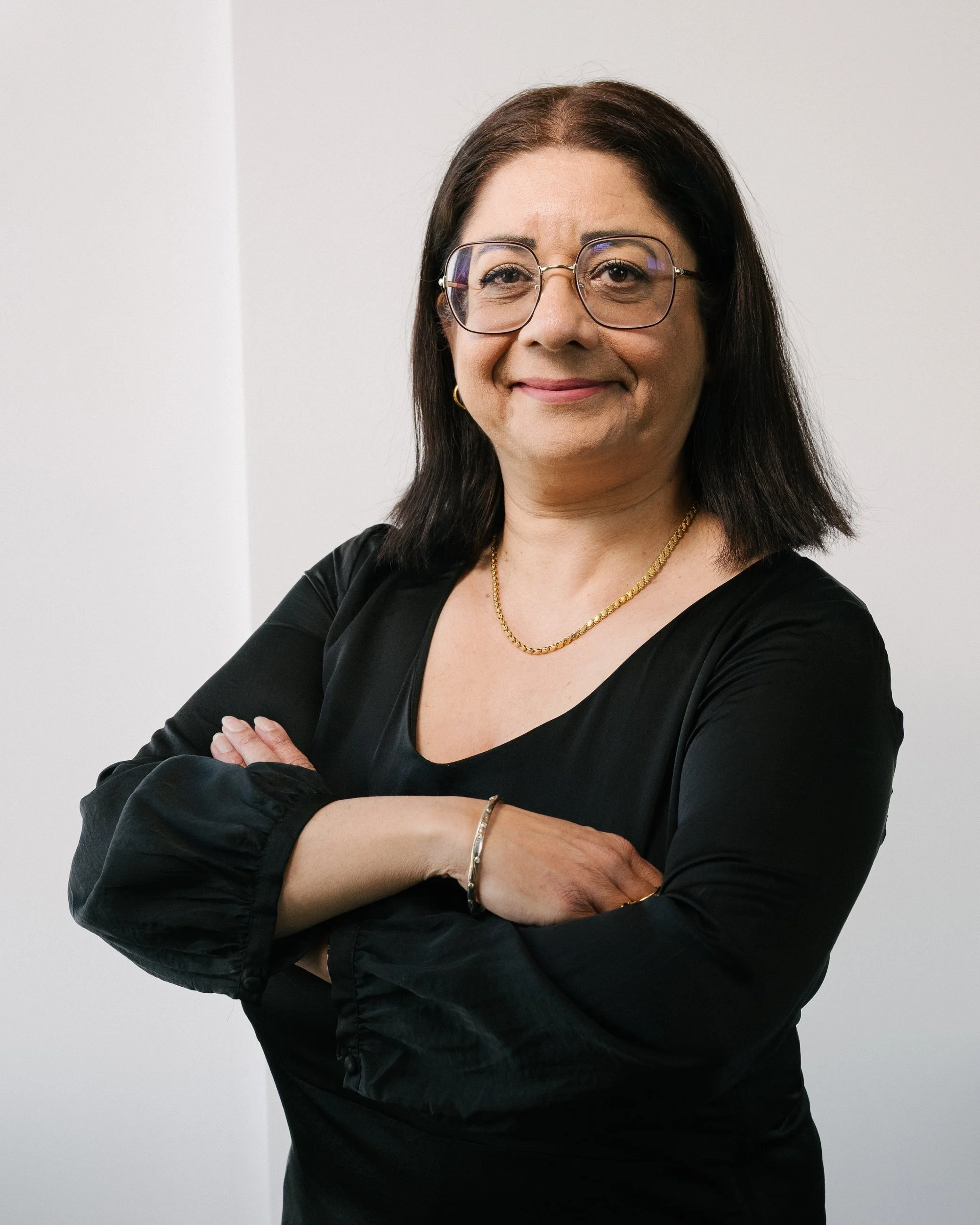 Porträt einer Frau mit dunklen Haaren, die eine Brille und ein schwarzes Oberteil trägt. Die Arme sind vor der Brust verschränkt. Hintergrund ist neutral.
