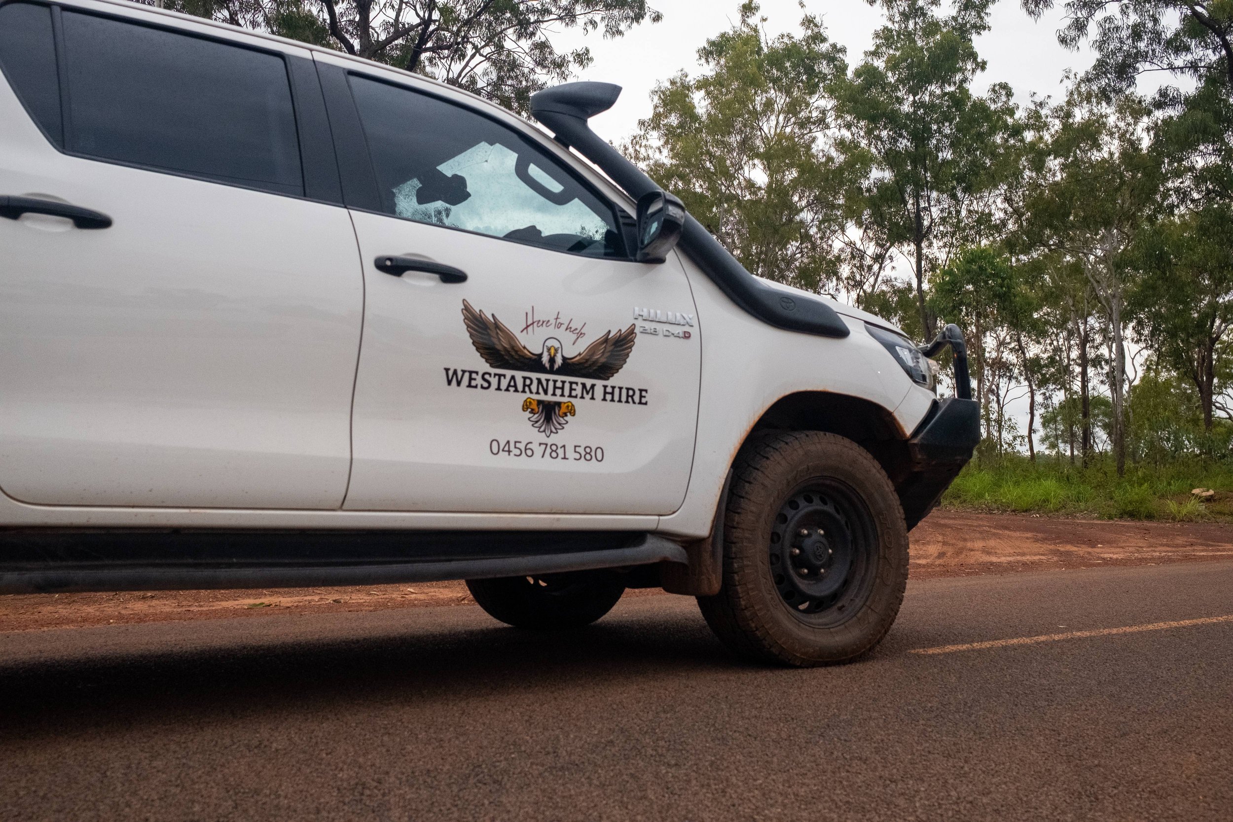White Toyota Hilux 4wd vehicle hired in Maningrida from "WESTARNHEM HIRE" logo parked on a road with trees in the background.