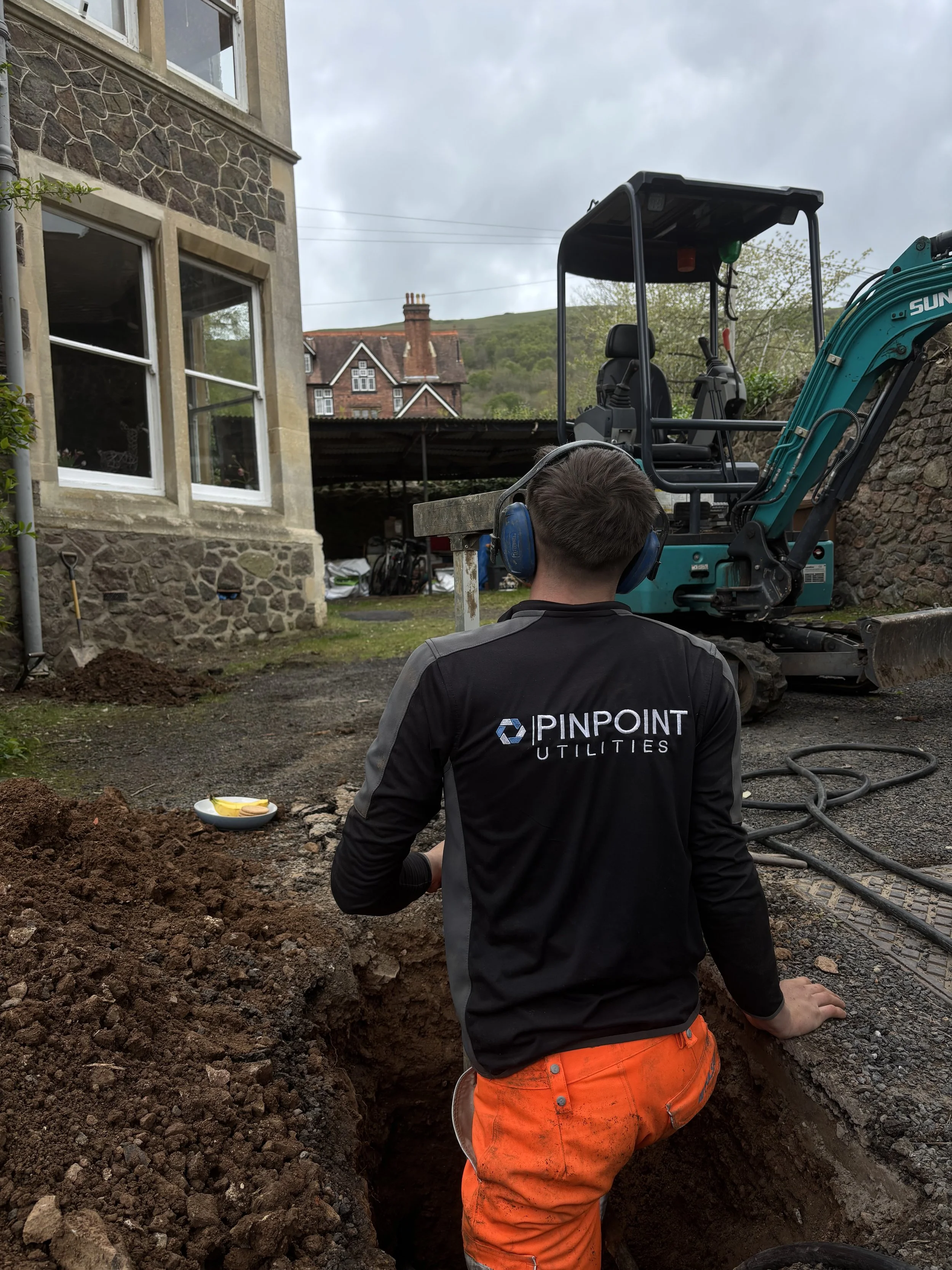 A worker with a black jacket with the logo PINPOINT UTILITIES, orange pants, and a headset is working in a deep trench outside a house with a stone facade, during cloudy weather. An excavator is nearby, and a multi-story house with a chimney and a hill can be seen in the background.