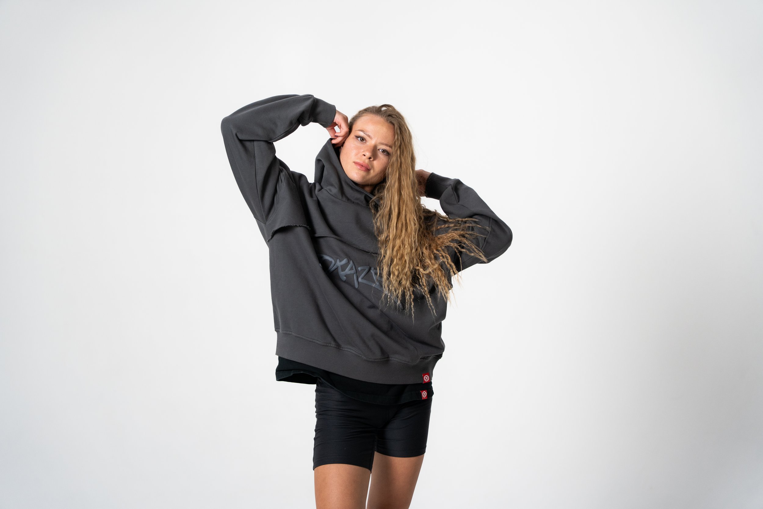 Person wearing a gray hoodie and black shorts, posing with hands near head against a plain white background.