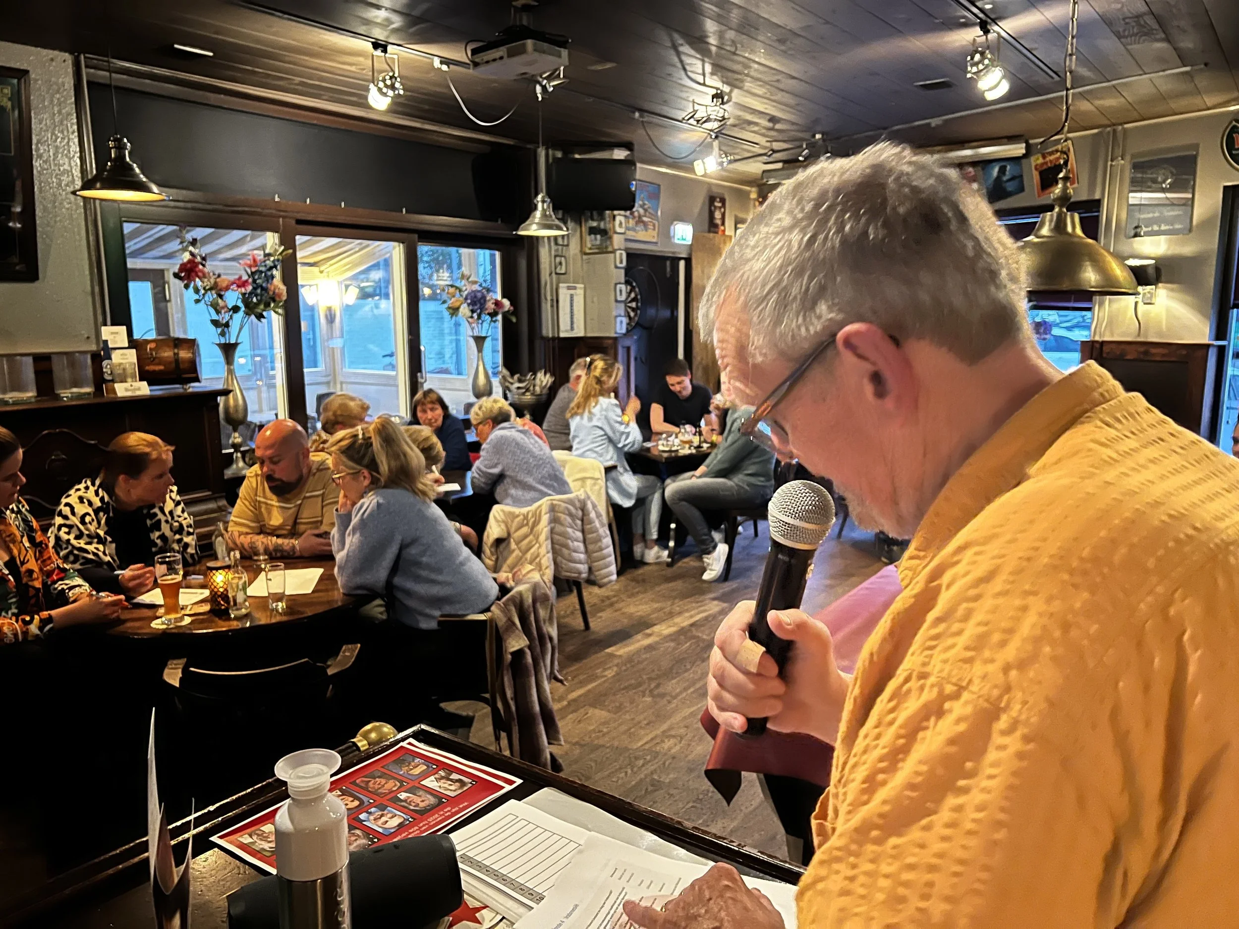 Een man met grijze haren en een geel overhemd leest of noteert iets terwijl hij een microfoon vasthoudt in een restaurant of café. Op de tafel voor hem liggen menu's, een fles en papieren.