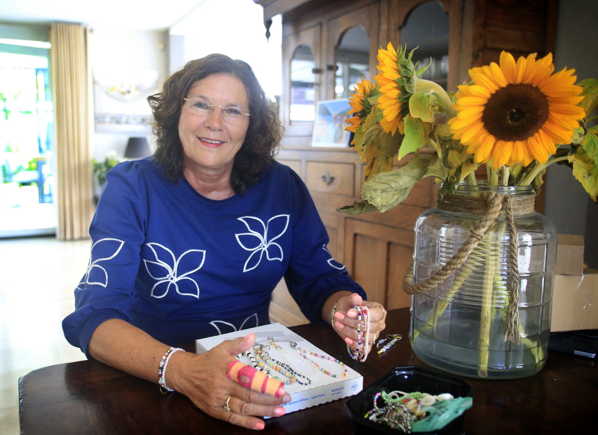 Vrouw zit aan een tafel met sieraden en een grote vaas met zonnebloemen, binnenruimte.
