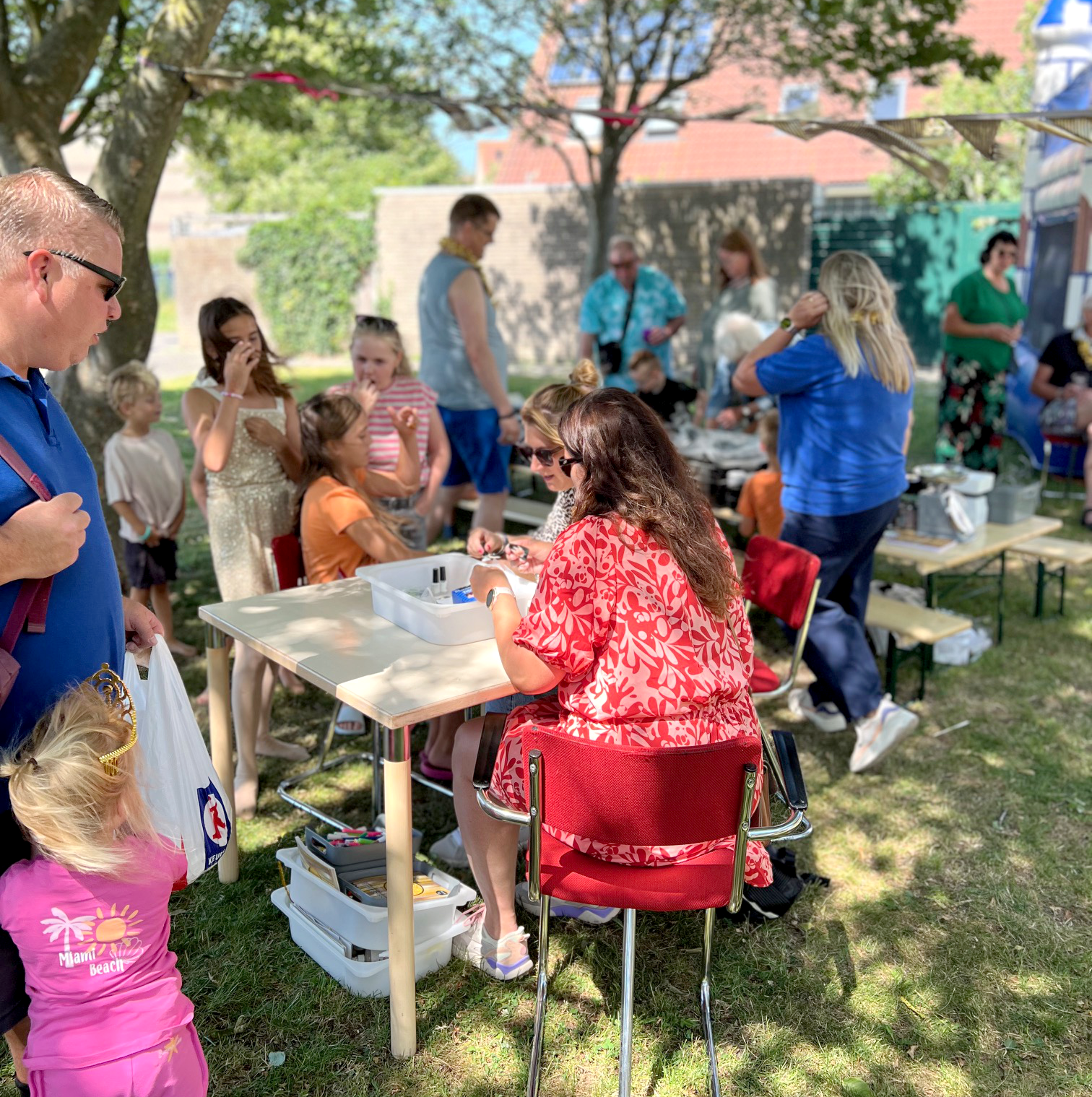 Mensen verzamelen zich bij een outdoor festival of markt in een tuin, met kinderen en volwassenen die genieten van activiteiten onder een boom en langs tafels.