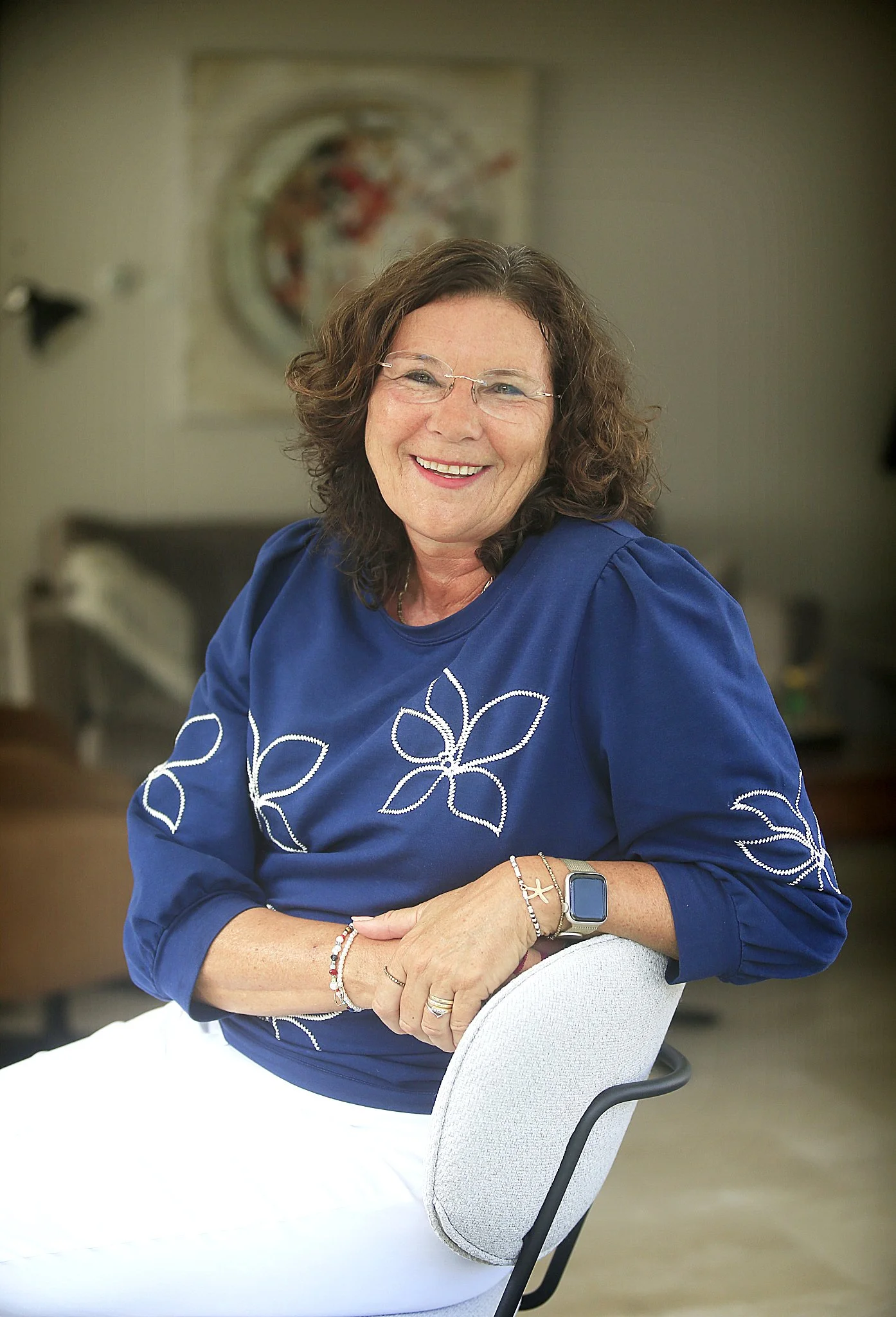 Portret van een lachende vrouw met bruin, krullend haar, zittend op een stoel, in een binnenruimte met moderne kunst op de muur op de achtergrond.