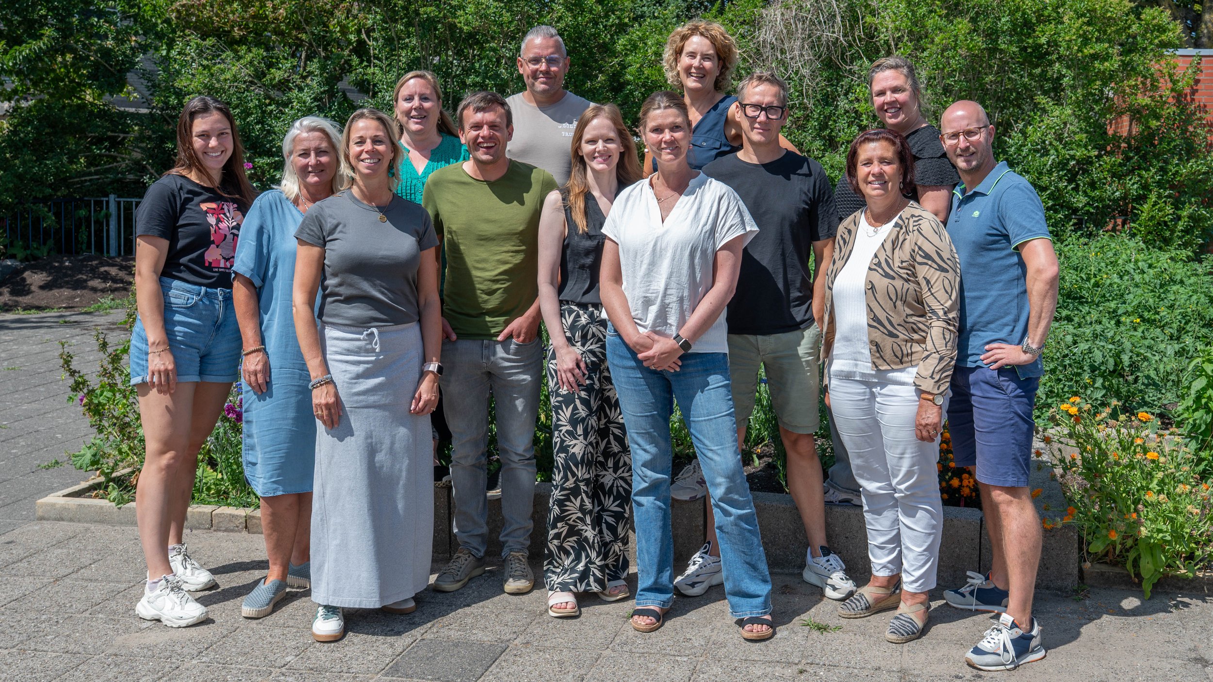Groep mensen poseren buiten op een zonnige dag, met groene struiken en bloemen op de achtergrond.