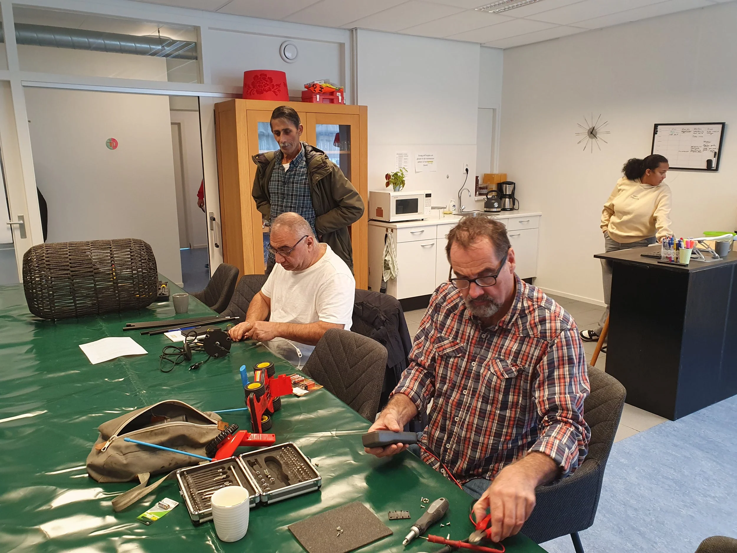 Mannen werkt met gereedschap en elektronische apparatuur op een tafel in een kantoorruimte, terwijl andere mensen in de achtergrond bezig zijn.