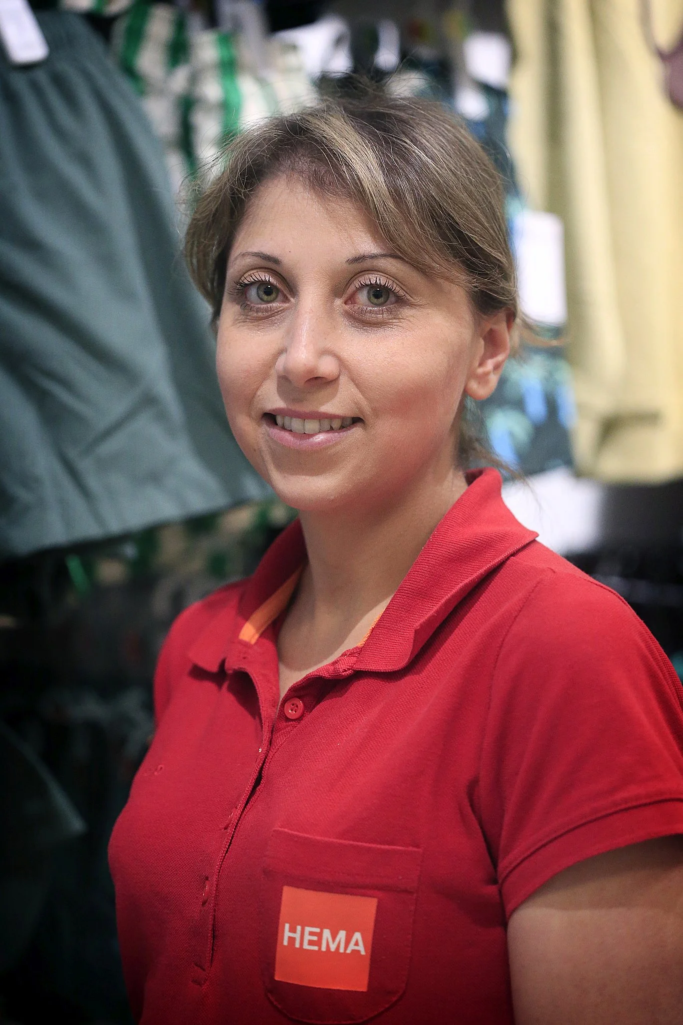 Vrouw in een rode HEMA-uniform poseert in een winkel, achtergrond met kleding.