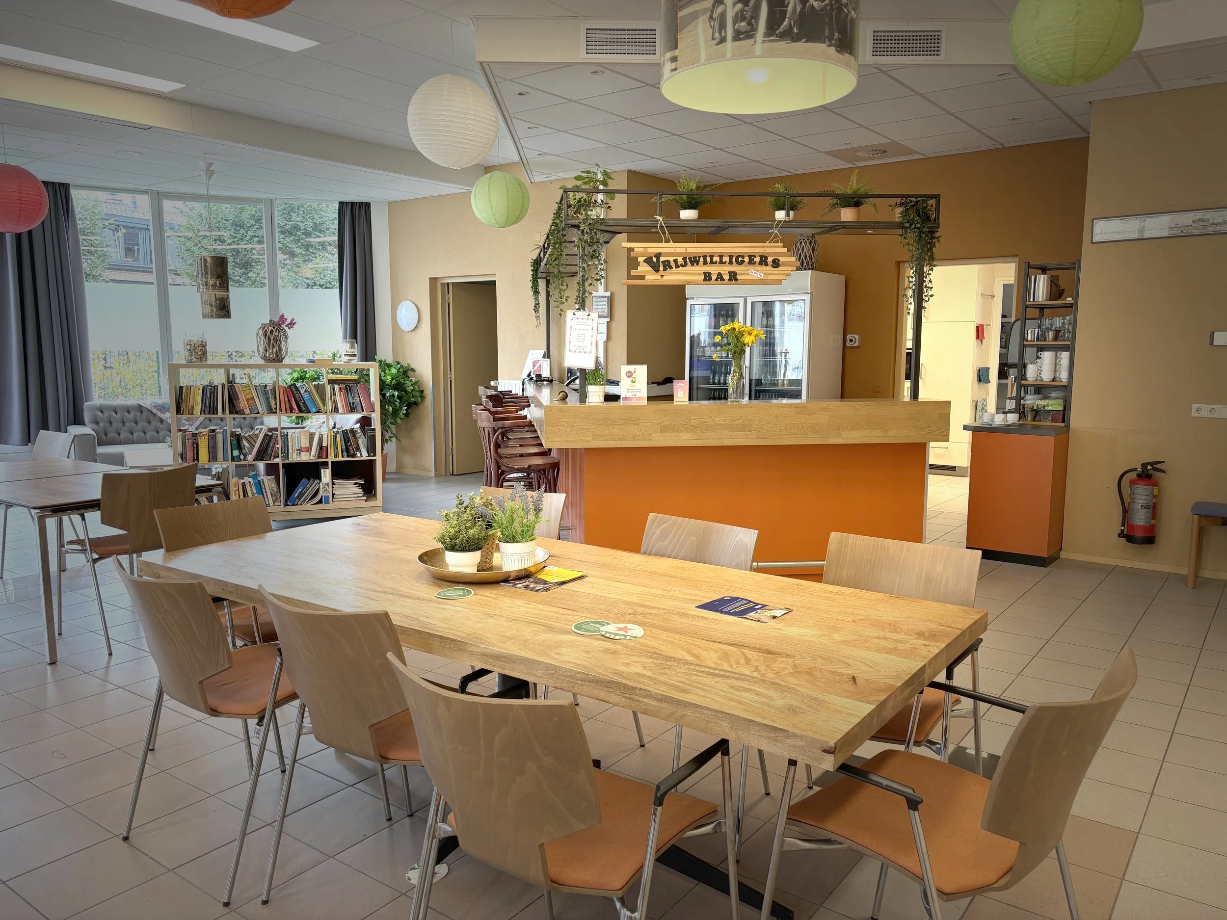 Binnenkant van een gezellige cafe of bibliotheek met een grote houten tafel, planten, boeken en een bar onder een bamboe plafond met hangende papieren lampen.