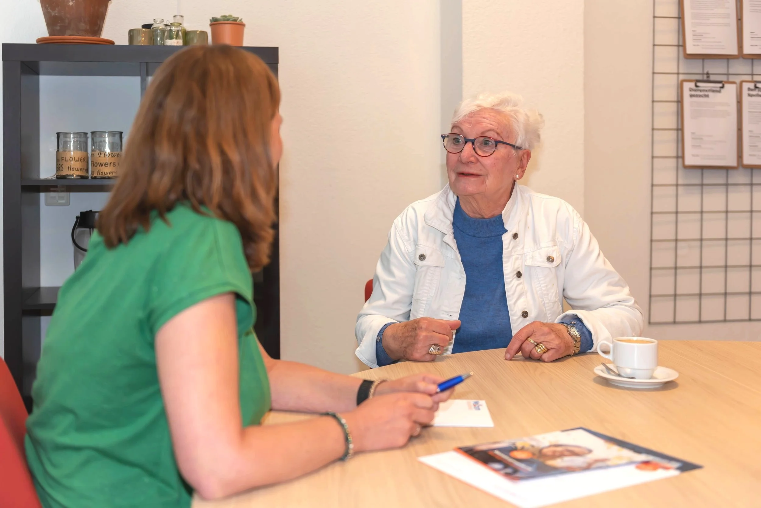 Twee vrouwen zitten aan een houten tafel in een bespreekruimte, ze hebben een gesprek. Een vrouw met bruin haar draagt een groene blouse, de andere vrouw heeft wit haar. Dit is een adviesgesprek.
