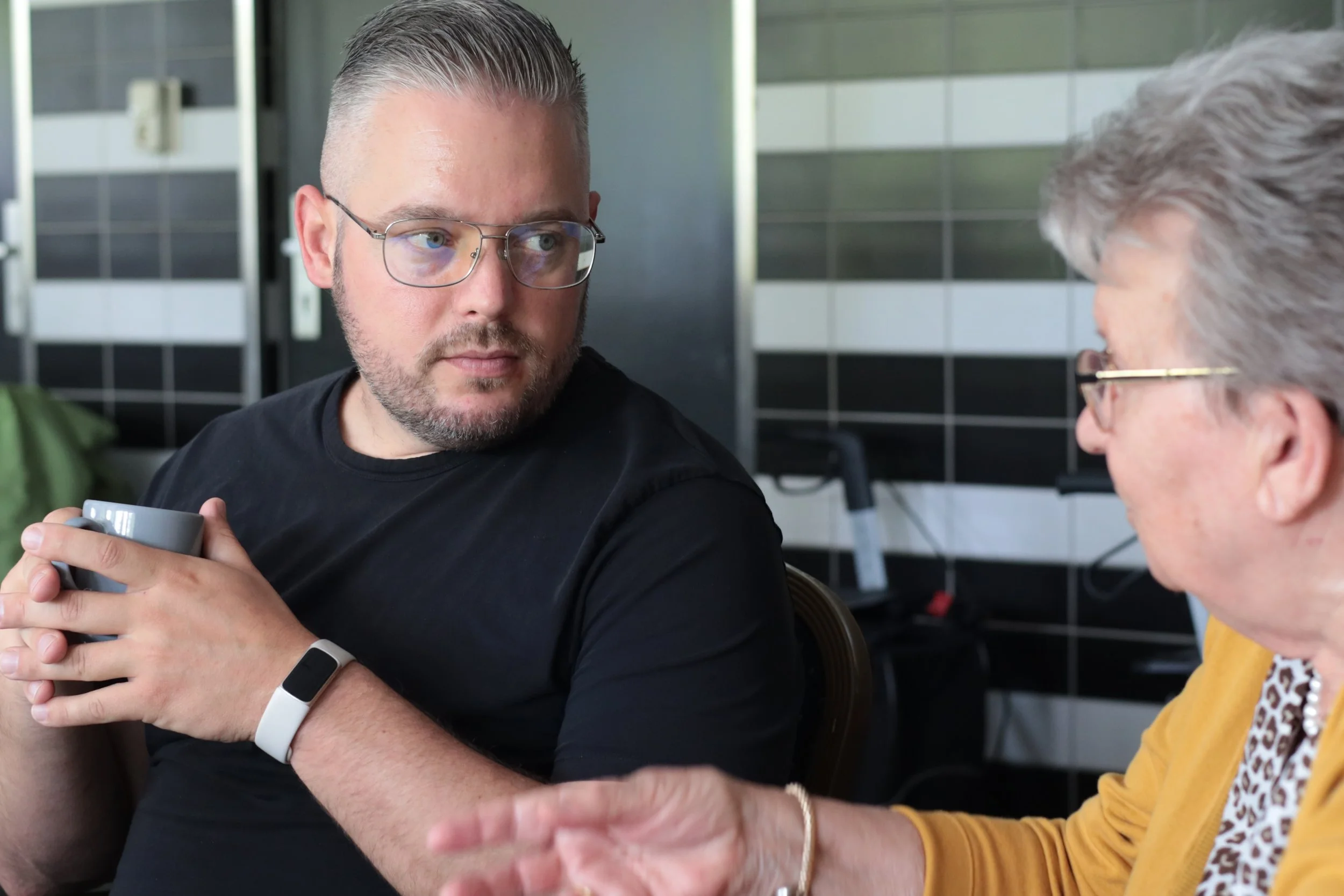 Roy van Schie houdt een mok en kijkt naar een oudere vrouw die met haar hand gebaren maakt, in een moderne kamer met zwarte en witte tegels op de muur.