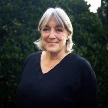 Vrouw met grijs haar en zwarte trui buiten met groene bomen op de achtergrond.