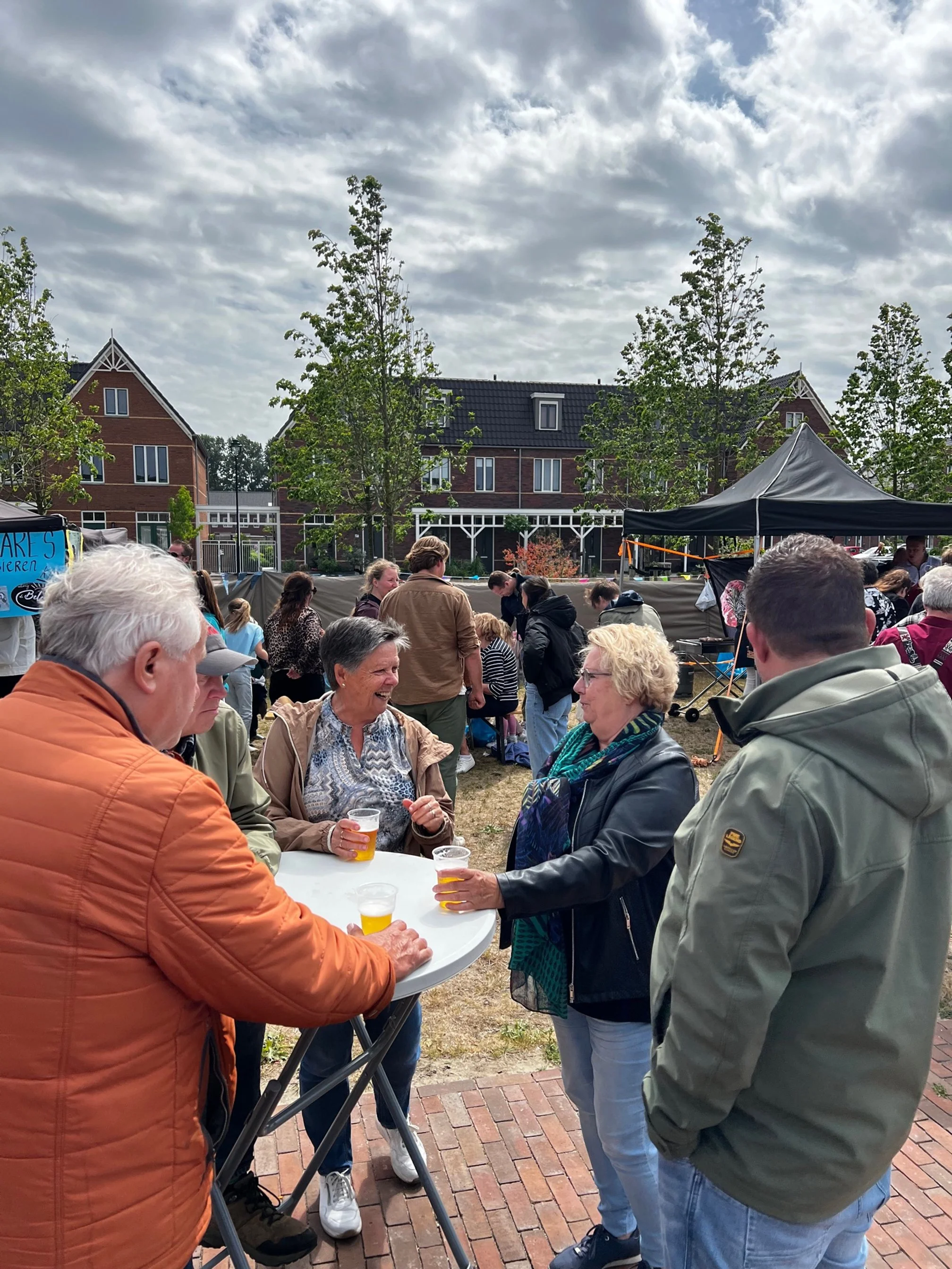 Mensen genieten van een drankje bij een buitenfestival of markt, onder een bewolkte hemel, met enkele bomen en huizen op de achtergrond.