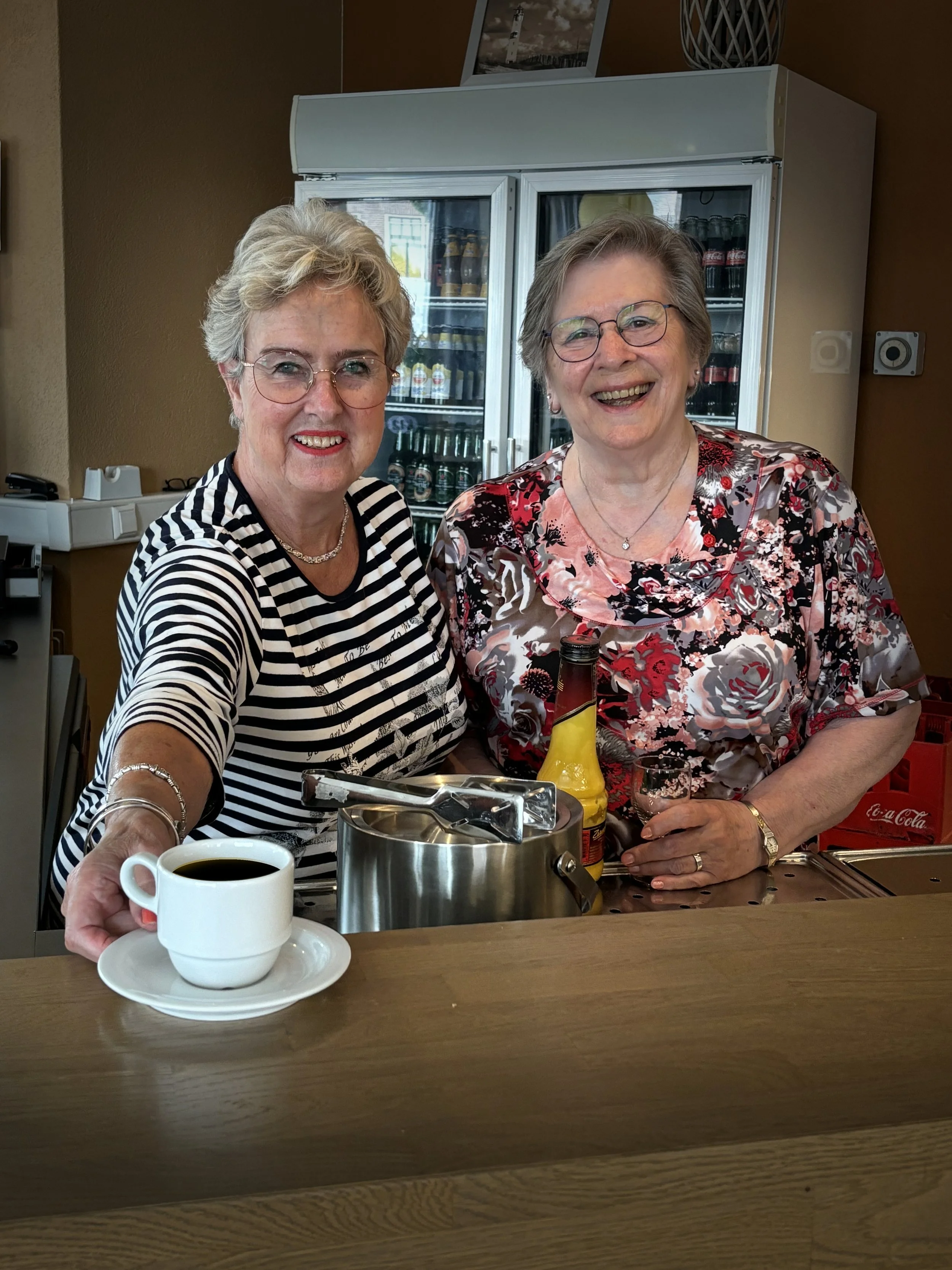 Twee oudere vrouwen lachen en drinken koffie achter de bar in een café of restaurant.