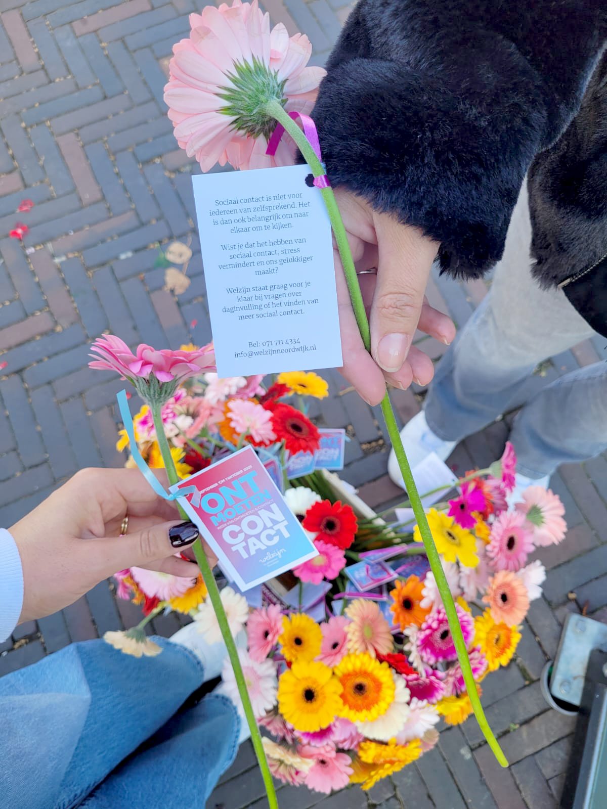 Twee mensen die een bloem met een kaart vasthouden. Op de grond staan bloemen in een emmer