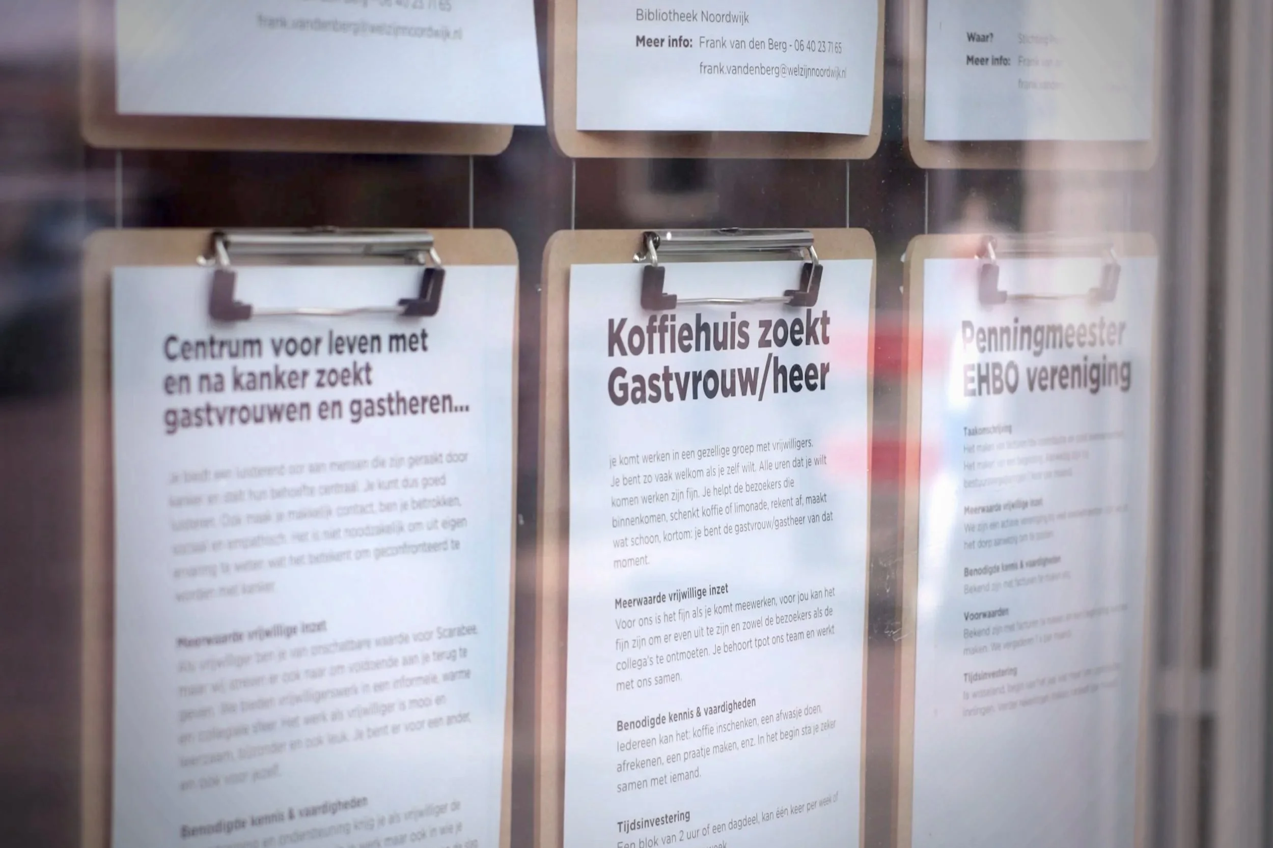Informatieborden met tekst over vrijwilligerswerk bij een koffietafel, op een glasraam. Tekst in het Nederlands, titels zoals 'Koffiehuis zoekt Gastvrouw/heer'.