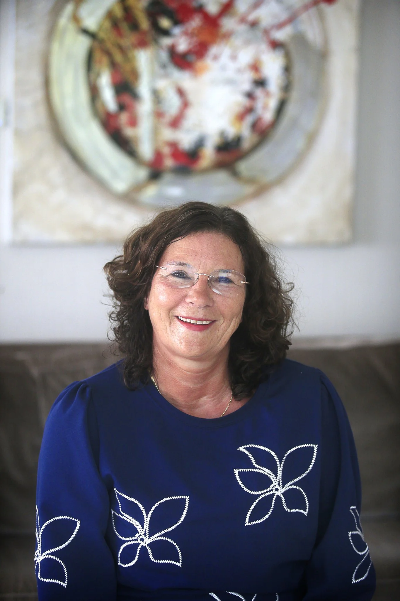 Portret van een lachende vrouw met bruin, golvend haar en bril, zittend op een bank met een abstract schilderij op de achtergrond.