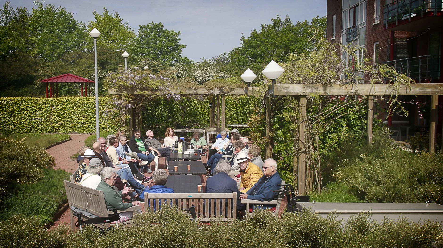 Een groep ouderen zit samen in een tuin op banken en stoelen, omringd door groen en bloemen. Ze lijken deel te nemen aan een sociale bijeenkomst, met enkele kopjes koffie en een tafel met apparatuur in het midden.