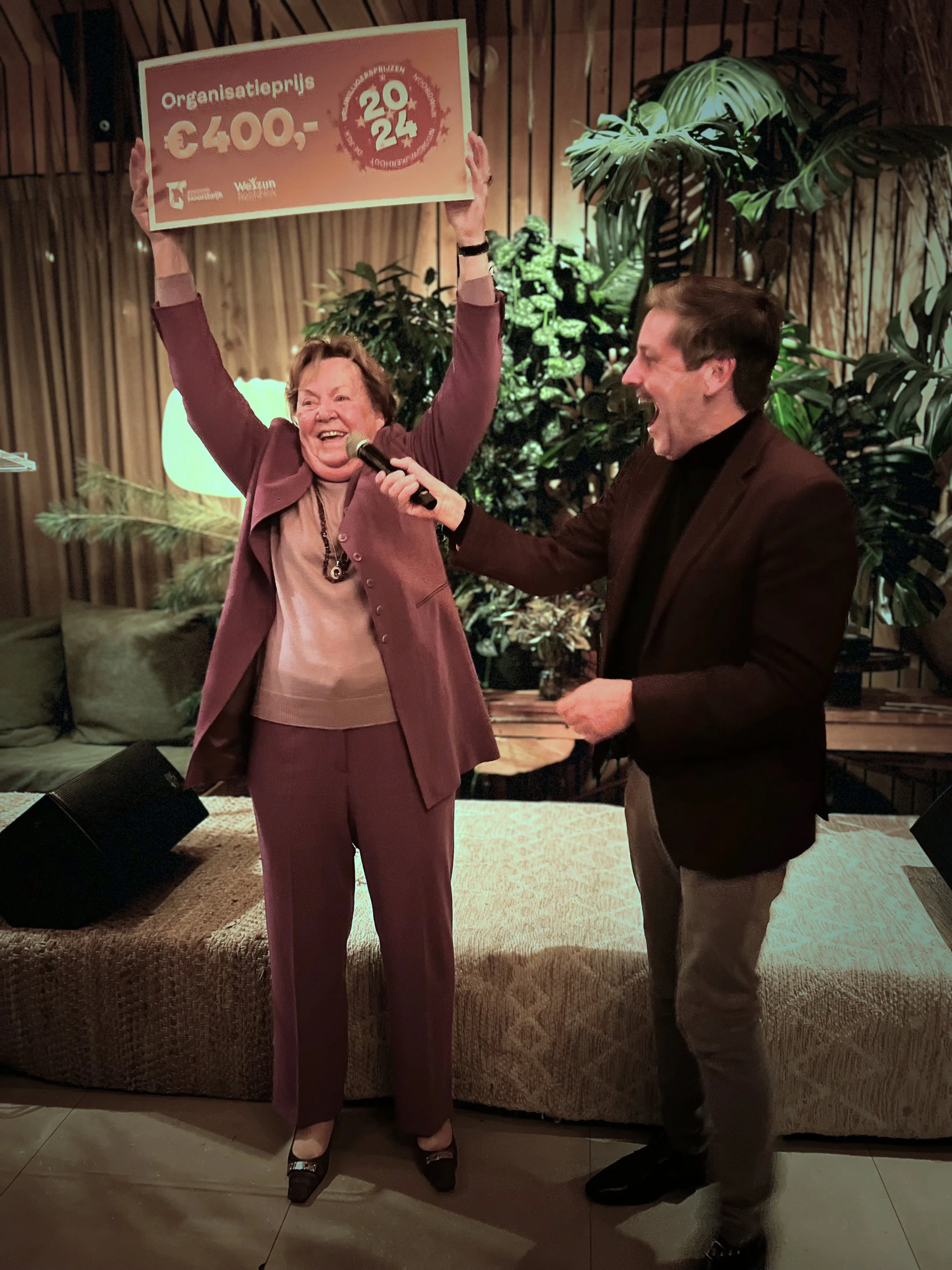 Een vrouw in een lila pak houdt een groot rood en wit cheque omhoog terwijl ze lacht, een man in een zwarte jas en grijze broek houdt een microfoon naar haar toe en lacht ook, in een gezellig interieur met groene planten op de achtergrond.