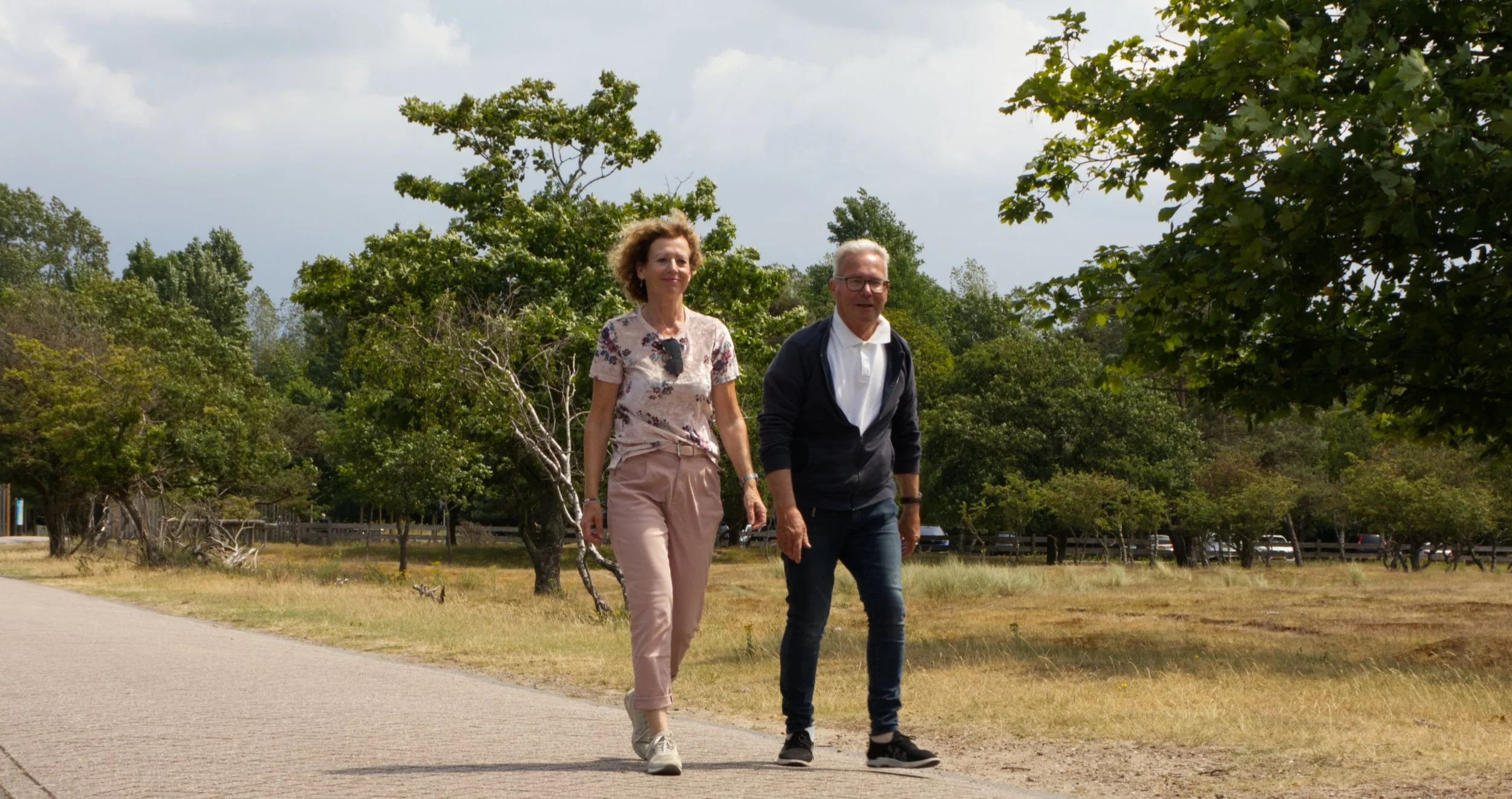 Twee mensen wandelen buiten op een pad, omgeven door bomen en gras, onder een bewolkte lucht.