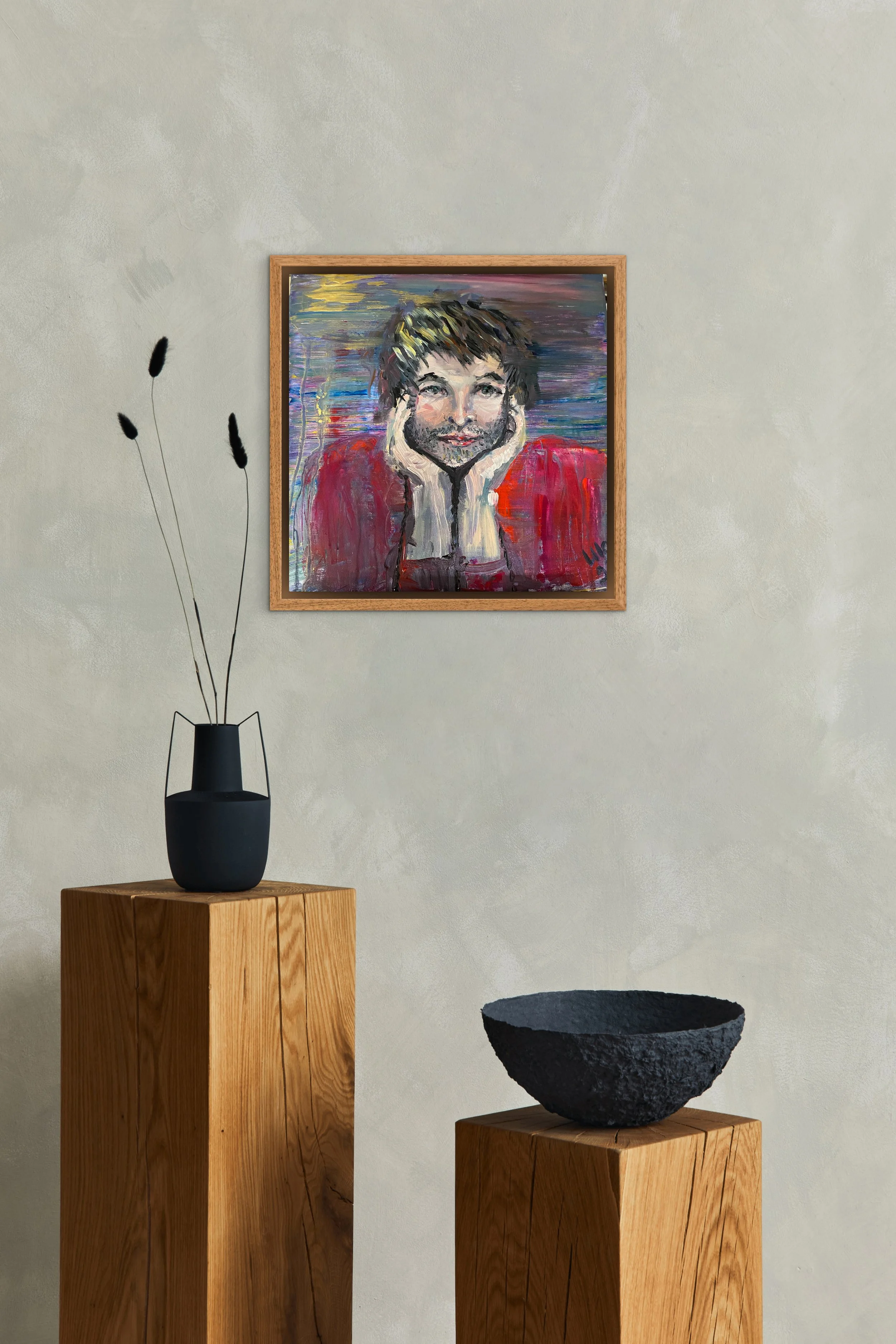 Ein Gemälde eines lächelnden Mannes mit braunen Haaren, der die Hände ans Gesicht hält, hängt an einer hellen Wand. Zwei Holzpodeste tragen eine schwarze Vase mit stilisierten Gräsern und eine schwarze, grob texturierte Schale.