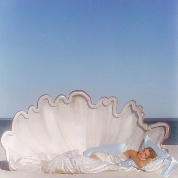 A person lying in a giant seashell bed on a beach, wrapped in a silky white sheet with pillows, under a clear blue sky.