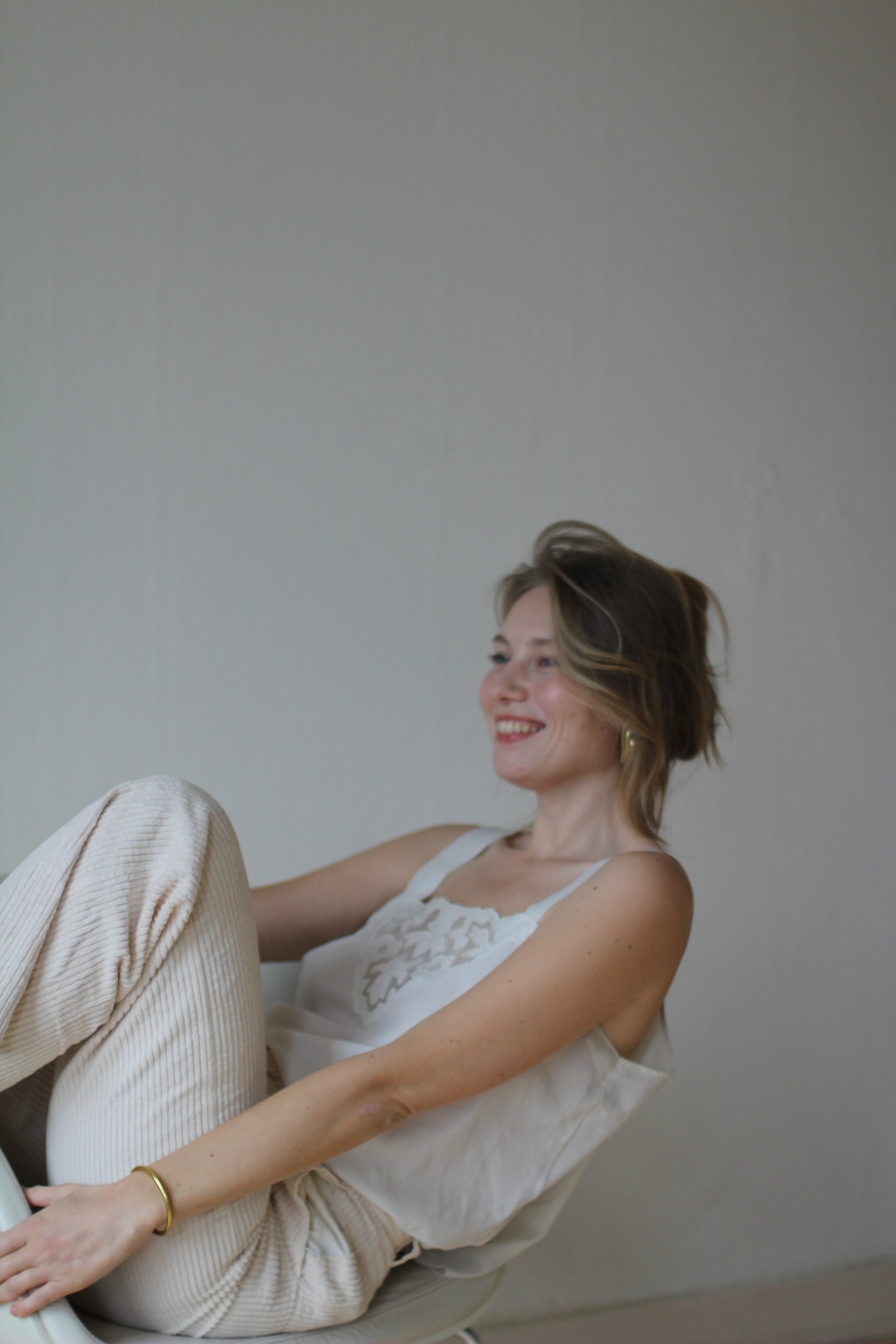 Smiling woman sitting in a chair, wearing a white top and light pants, with a relaxed pose.