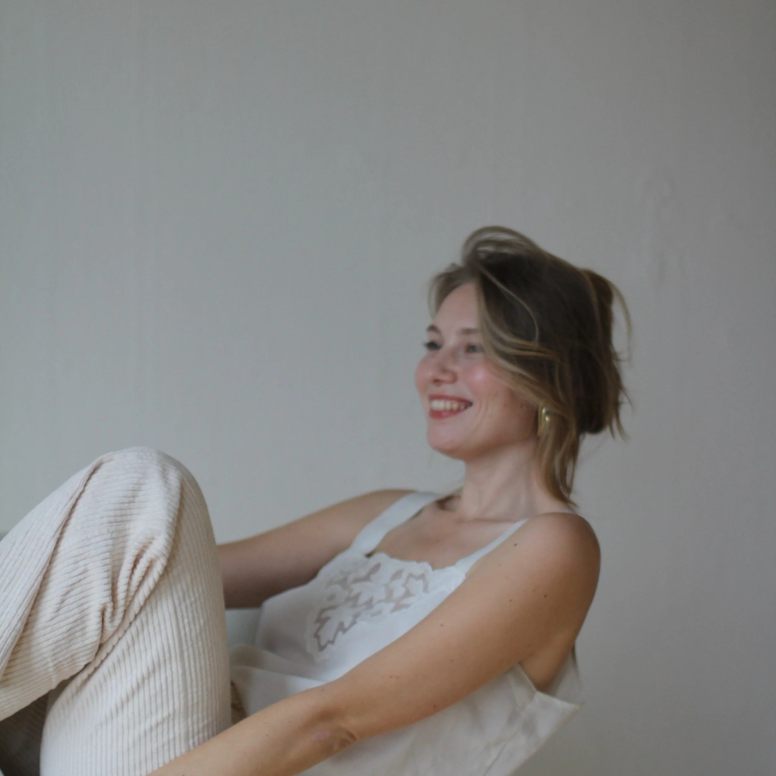 Woman sitting and smiling against a neutral background, wearing a light-colored top and pants.