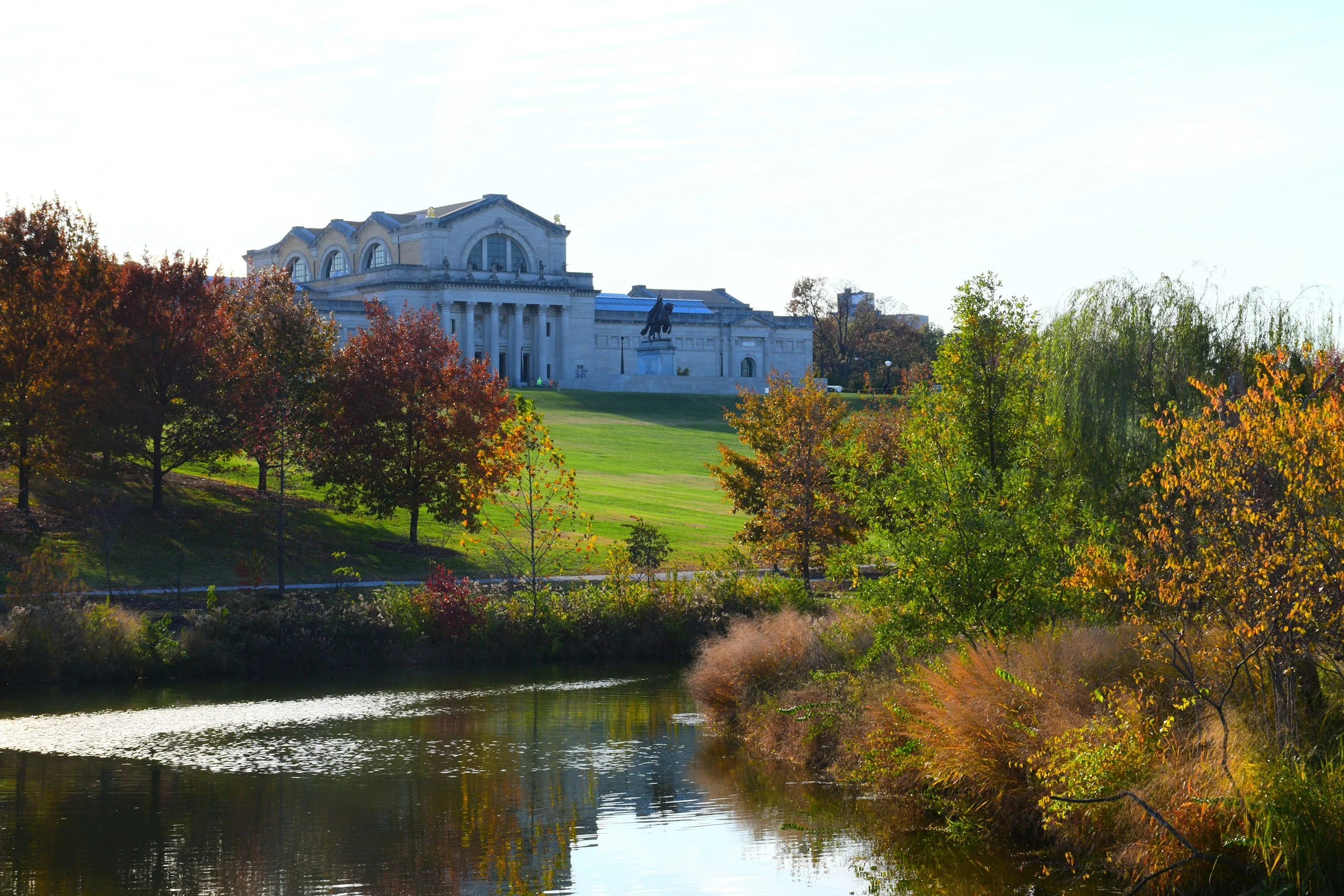 View of a grand marble building on a hill with columns and statues, surrounded by colorful autumn trees and a calm river reflecting the scenery.