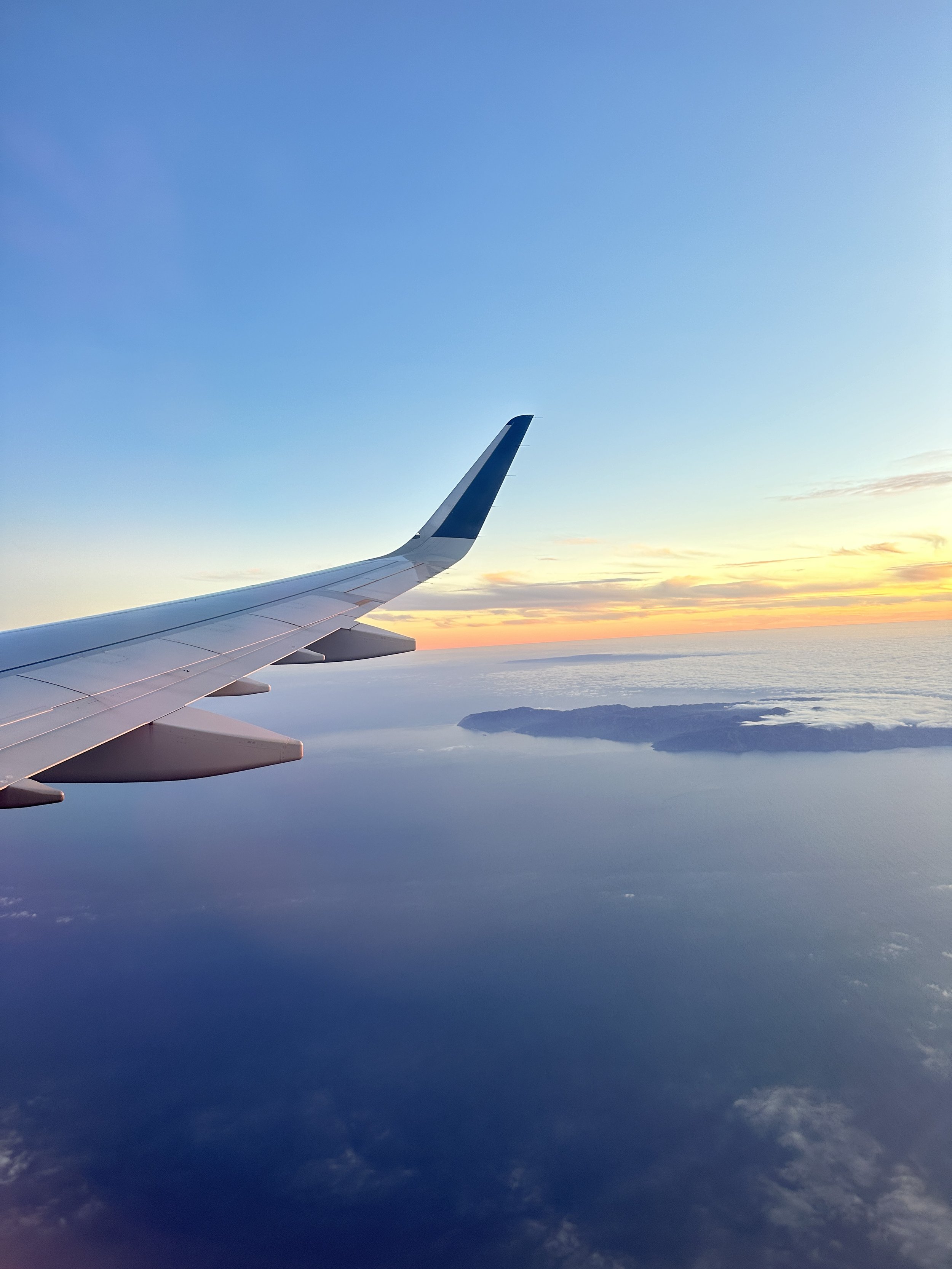 Airplane wing flying over ocean with island and sunset
