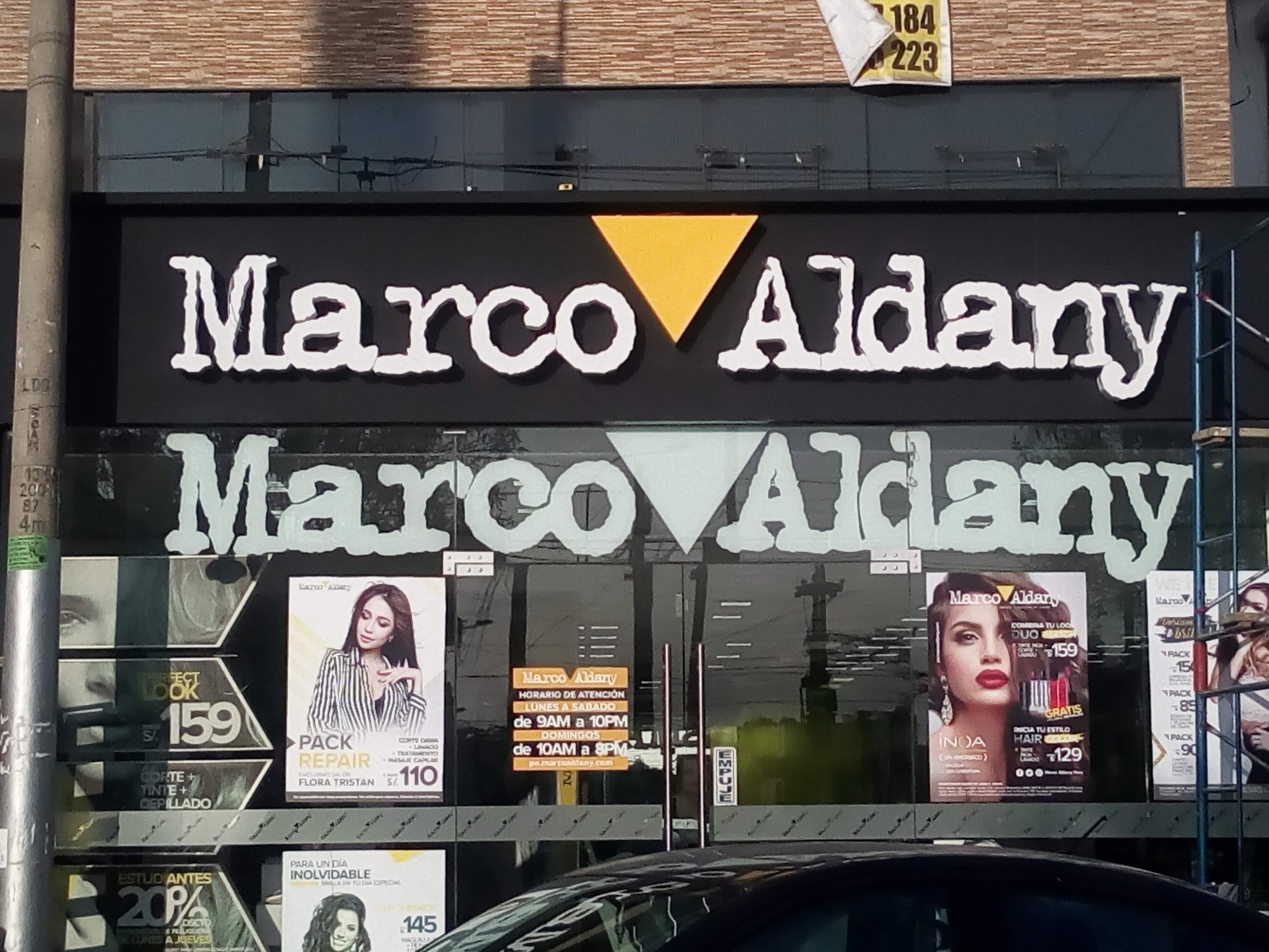 Fachada de una peluquería Marco Aldany con carteles promocionales de servicios de belleza. Hay posters con imágenes de mujeres mostrando diferentes estilos de peinado.