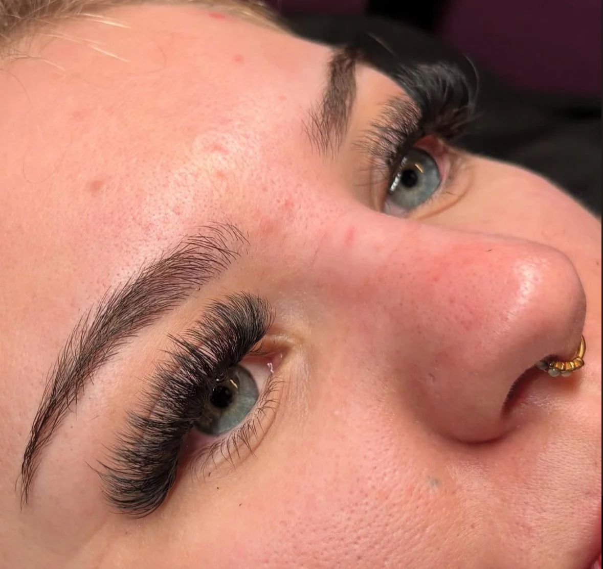 Close-up of a woman's face focusing on her eye with freshly done eyelash extensions, showing well-groomed eyebrows, light skin with some blemishes, a nose piercing, and minimal makeup.