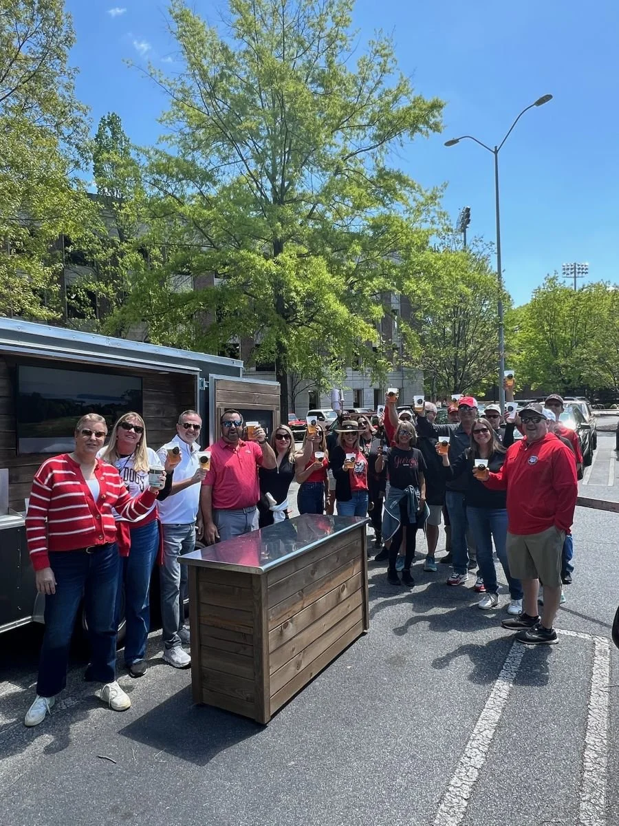 A group of people standing in front of Athens On Tap's mobile bar trailer for a tailgate outdoors, holding drinks, with trees and a clear blue sky in the background.