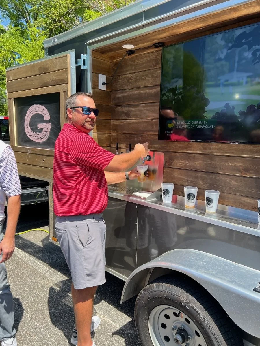 Guest pouring a drink from a tap on our wooden mobile bar with a TV screen, during an outdoor tailgate for a sporting event.