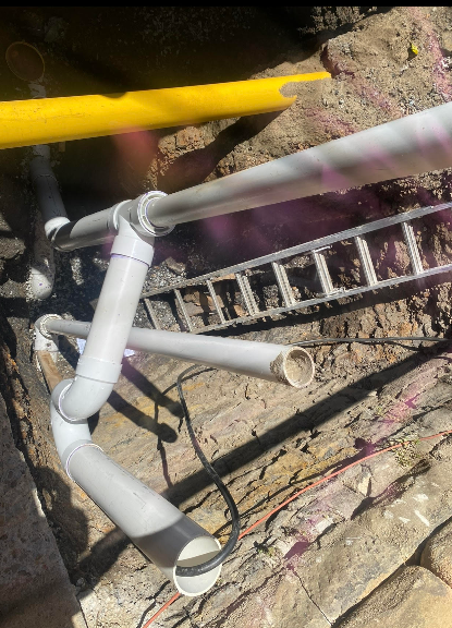 View of plumbing pipes and a metal ladder resting on a construction site with dirt ground and a yellow safety barrier.