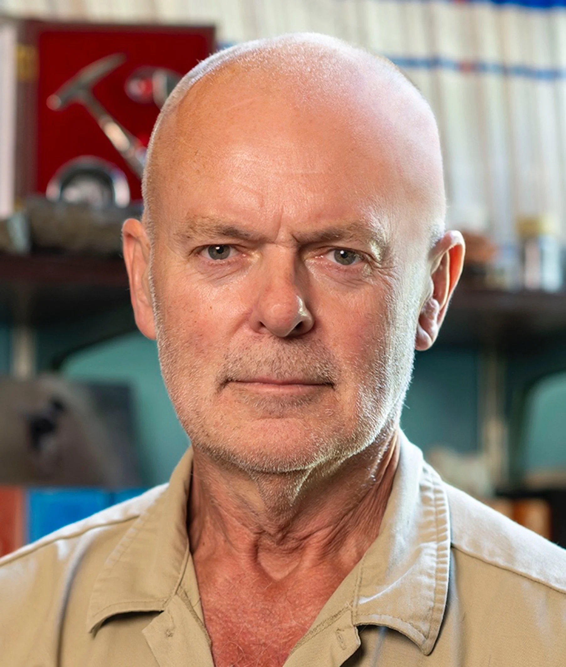Close-up portrait of an older man with a bald head and gray beard, wearing a beige shirt, in an indoor setting with blurred books and picture frames in the background.