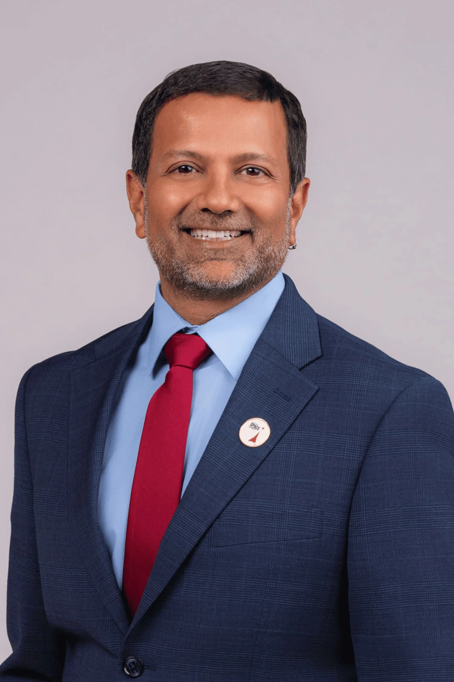 Professional man wearing a dark blue suit, light blue shirt, red tie, and a badge that says 'SPACE' with a rocket icon, smiling against a plain gray background.