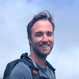 A smiling man with short dark hair, a beard, wearing a blue shirt and a backpack, outdoors with a cloudy sky in the background.