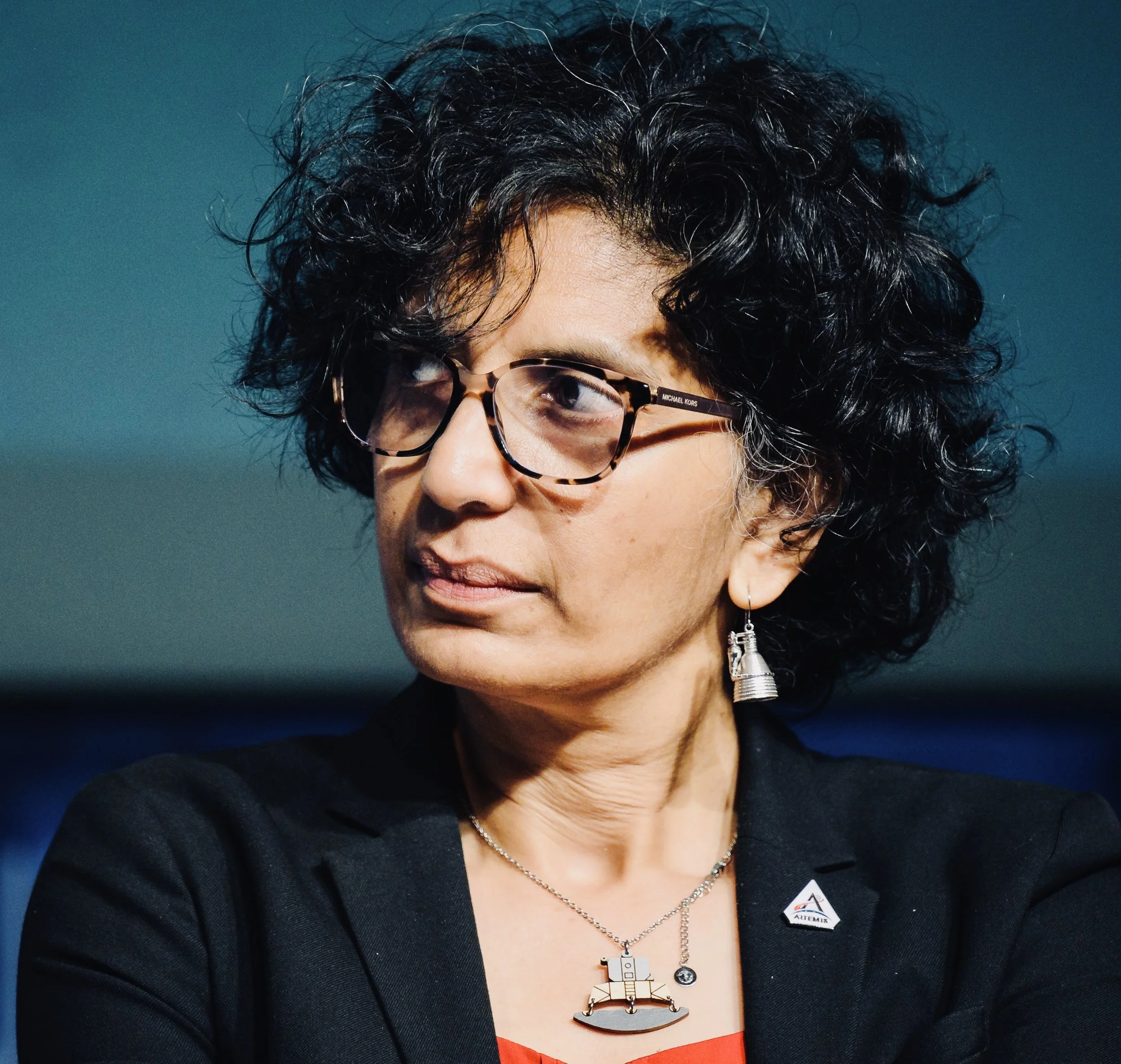 A woman with short curly black hair and glasses, wearing a black blazer with a NASA pin, a necklace with a space shuttle charm, and earrings, looking to her left against a blue-green background.