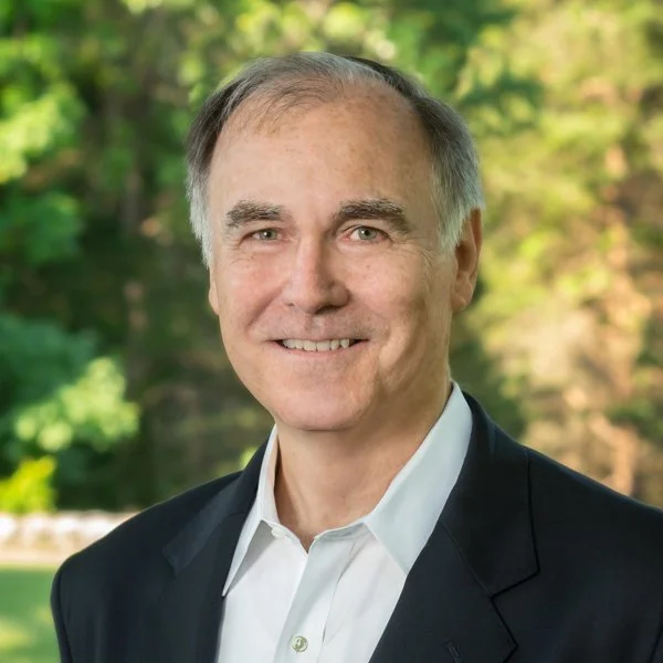 A middle-aged man with gray hair, wearing a dark suit jacket and white collared shirt, smiling outdoors with trees and greenery in the background.
