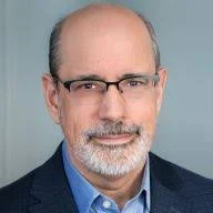 A portrait of a middle-aged man with short gray hair, a beard, glasses, wearing a suit jacket and blue shirt, against a gray background.