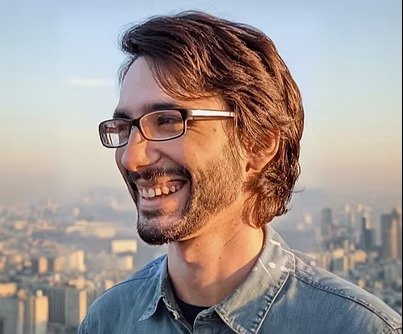 A man with glasses and a beard smiling outdoors with a city skyline in the background during sunset.