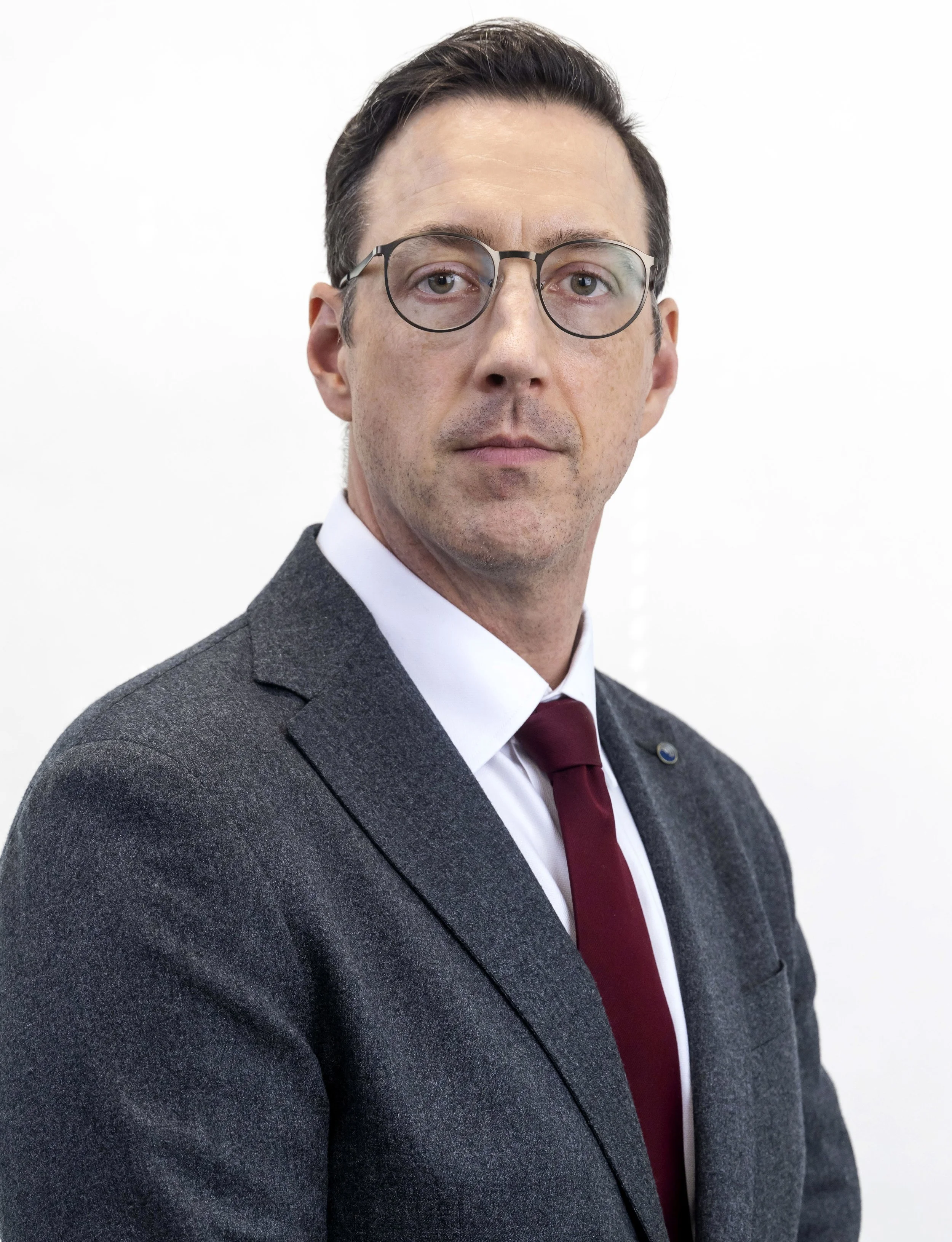 Portrait of a man in a suit with glasses, white shirt, and maroon tie against a plain white background.