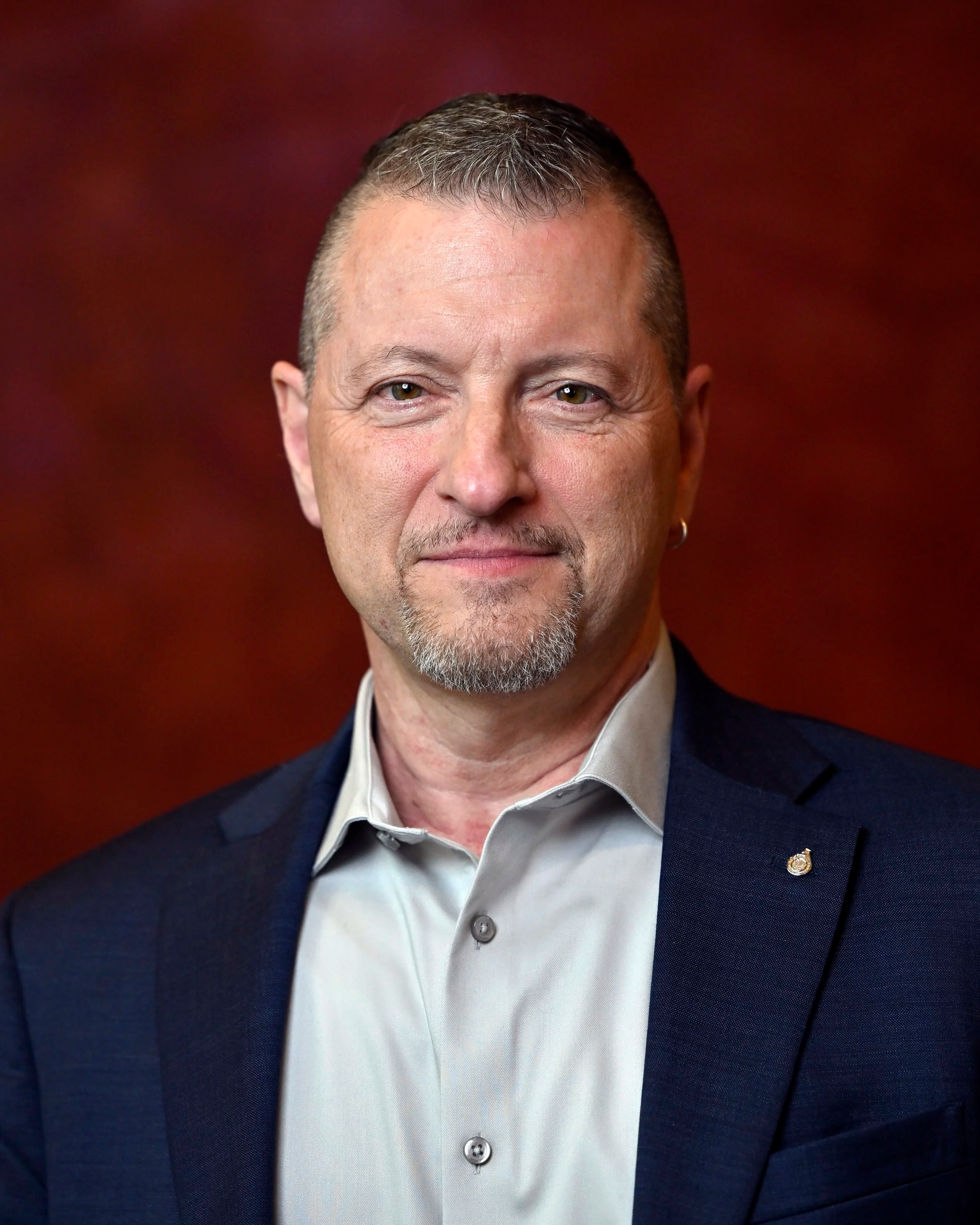 A middle-aged man with short, gray hair and a goatee wearing a dark suit jacket over a light-colored shirt, facing forward with a slight smile, against a blurred red background.