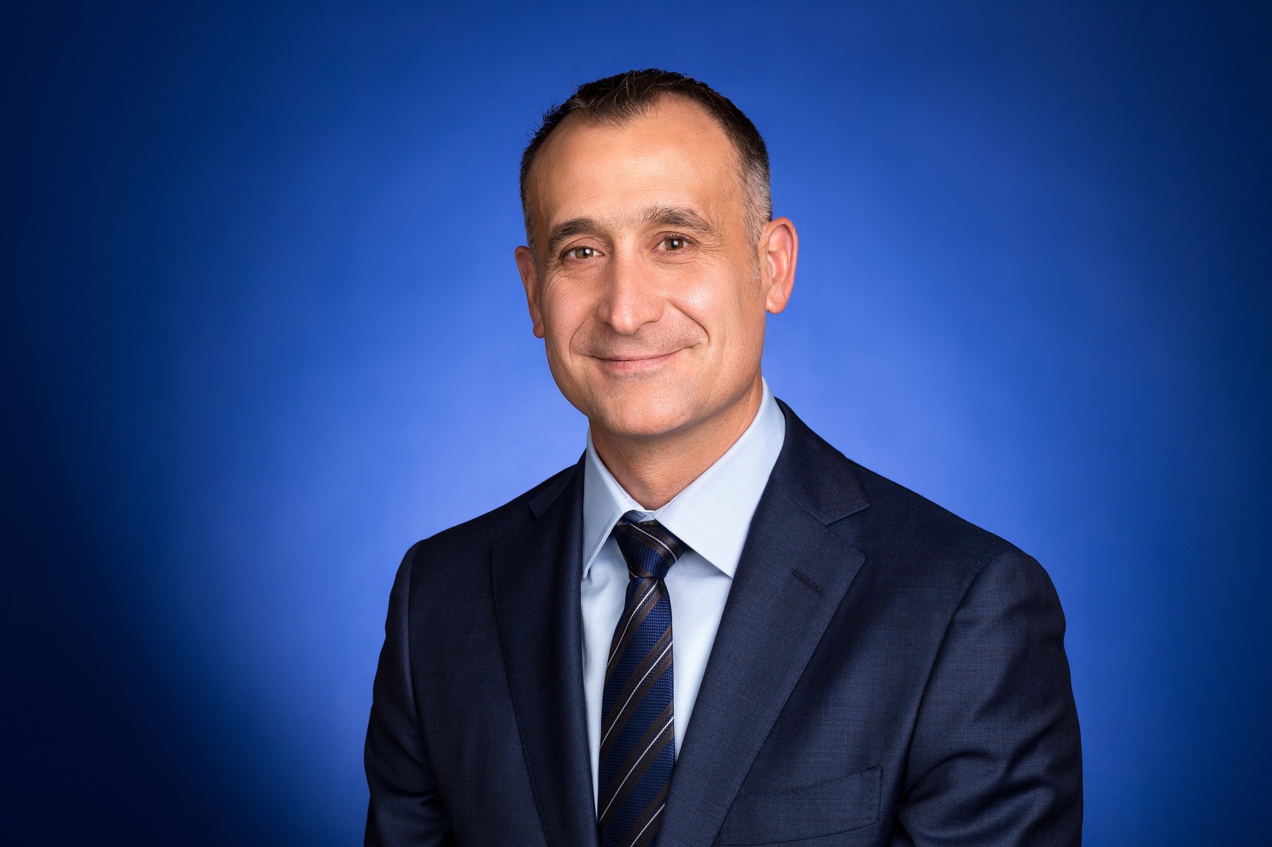 A professional man wearing a navy suit, light blue shirt, and a striped tie, smiling at the camera against a solid blue background.