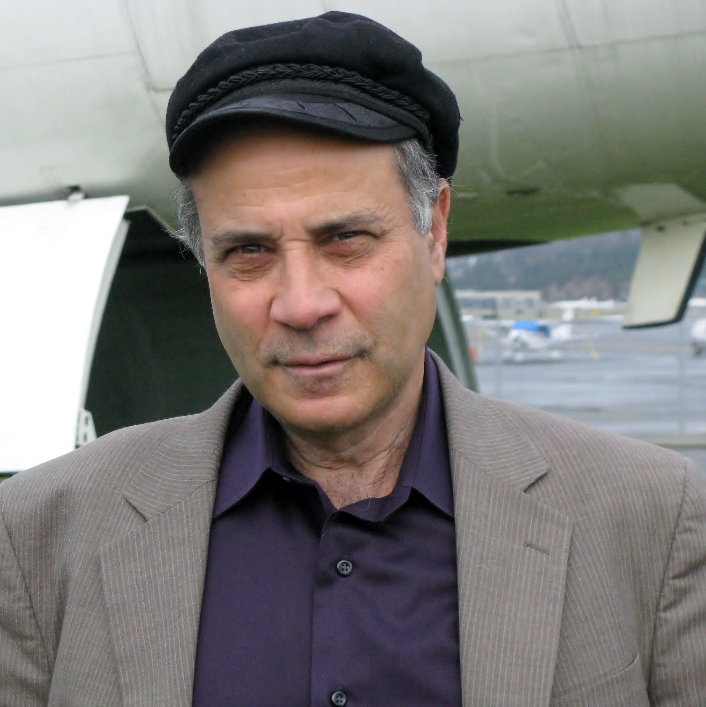 A middle-aged man wearing a black hat, dark purple shirt, and beige blazer standing near an airplane on an airport tarmac.