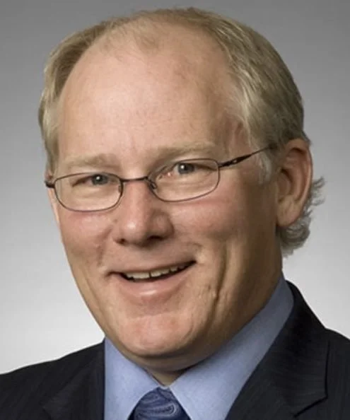 A middle-aged man with glasses, light-colored hair, and a friendly smile, wearing a dark suit jacket and a blue shirt against a neutral background.
