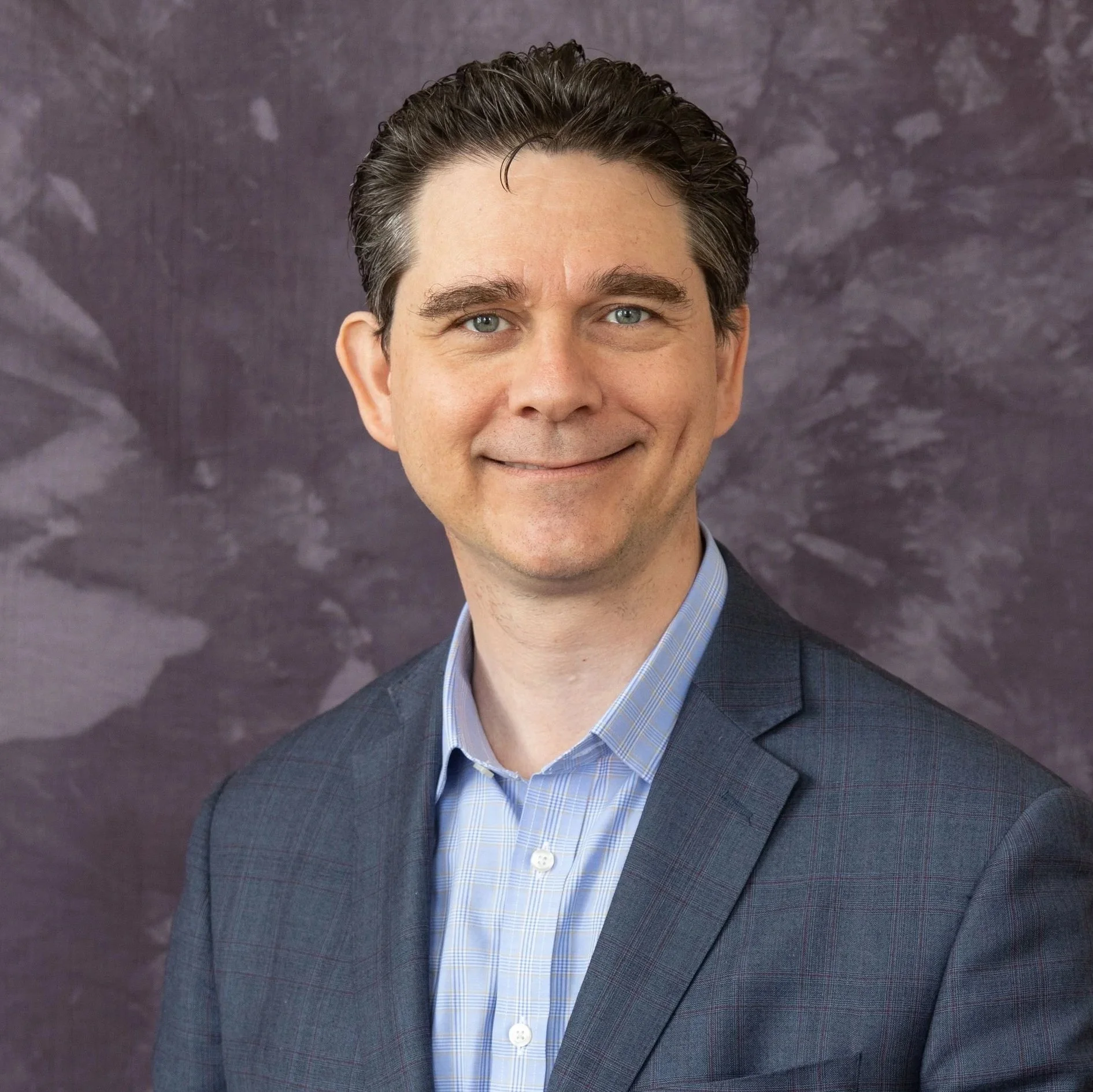 A man in a suit and light blue dress shirt, smiling in front of a textured dark background.