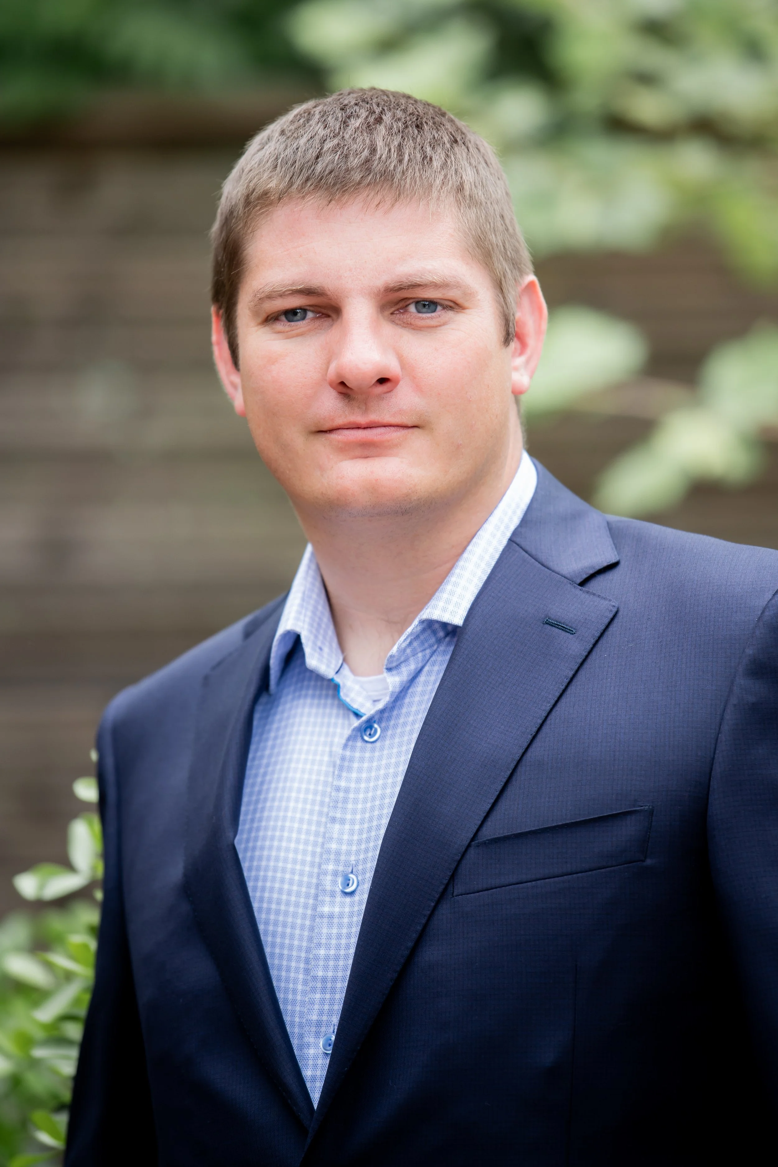 A man in a blue suit and light blue checkered shirt standing outdoors with a wooden fence and greenery in the background.